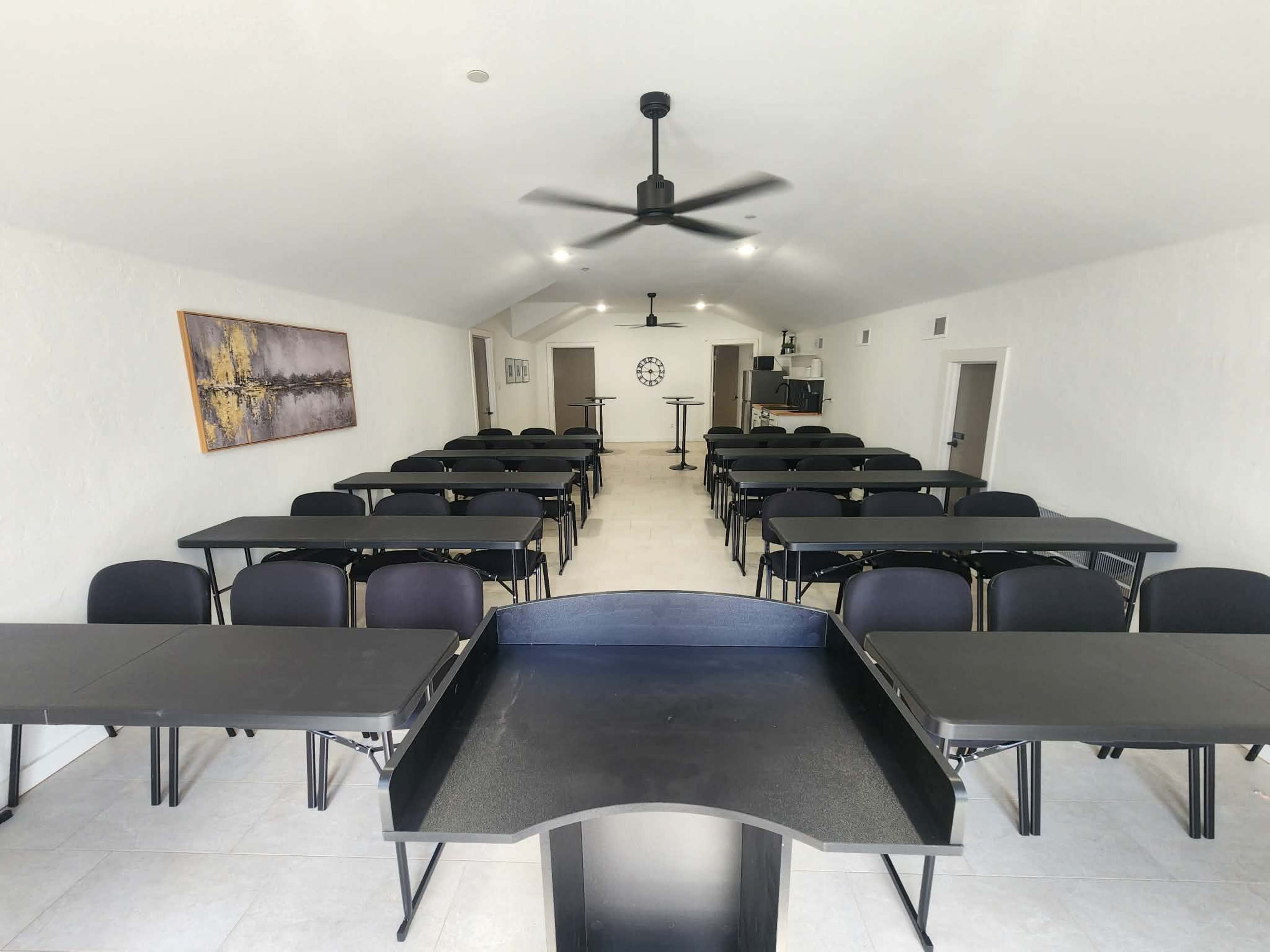 The image shows a large, empty classroom arranged with rows of black tables and chairs, featuring a podium at the front and a clock on the wall.