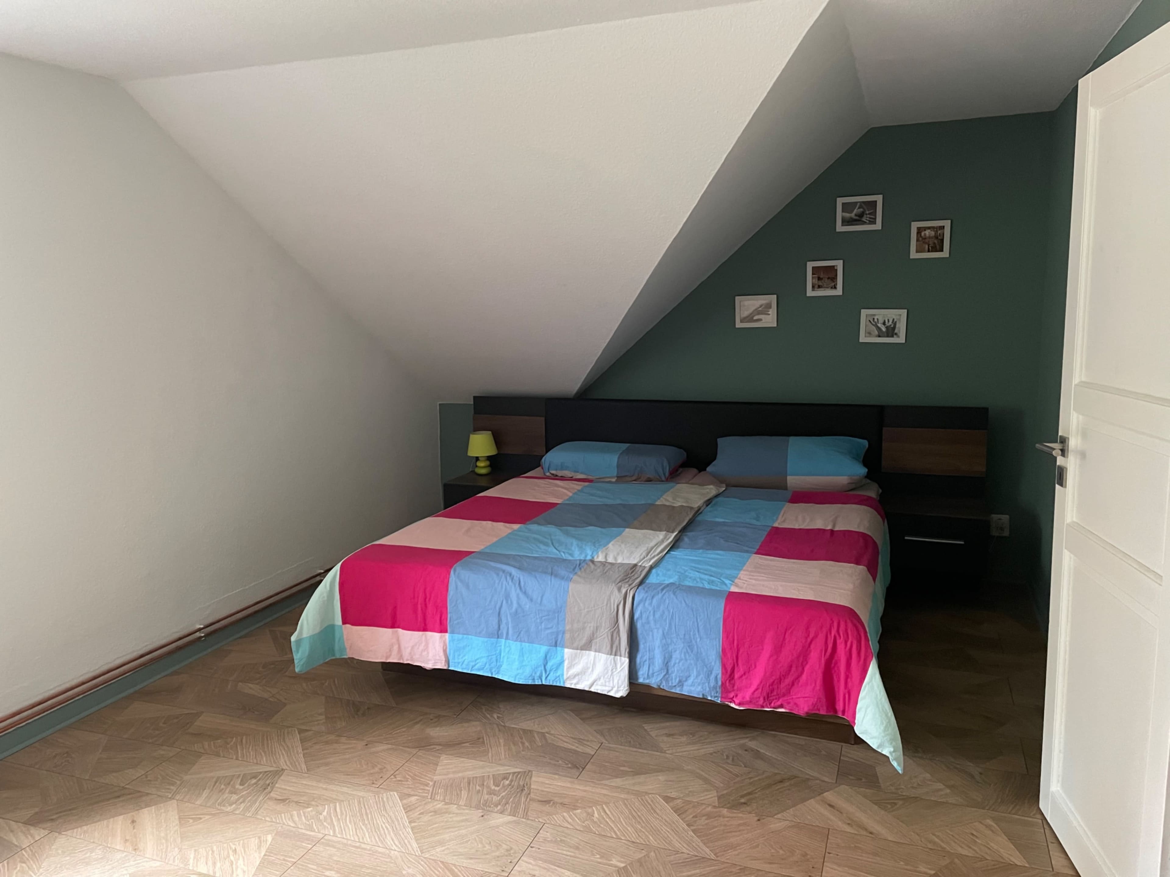 A bedroom with a sloped ceiling, featuring a double bed dressed in a colorful patchwork blanket and a small green lamp on a bedside table.