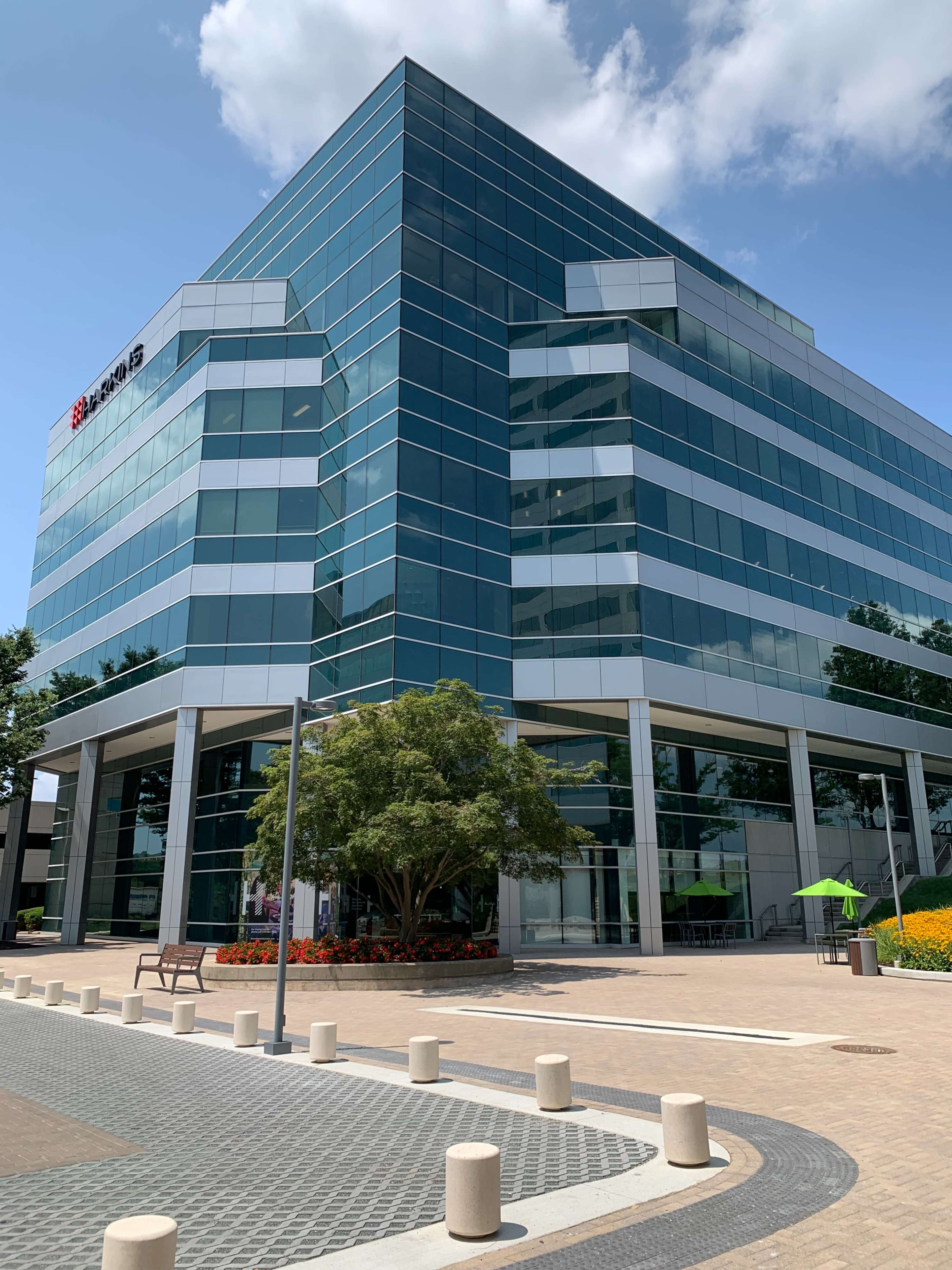 The image shows a modern glass office building with multiple stories, surrounded by landscaping and a paved walkway.