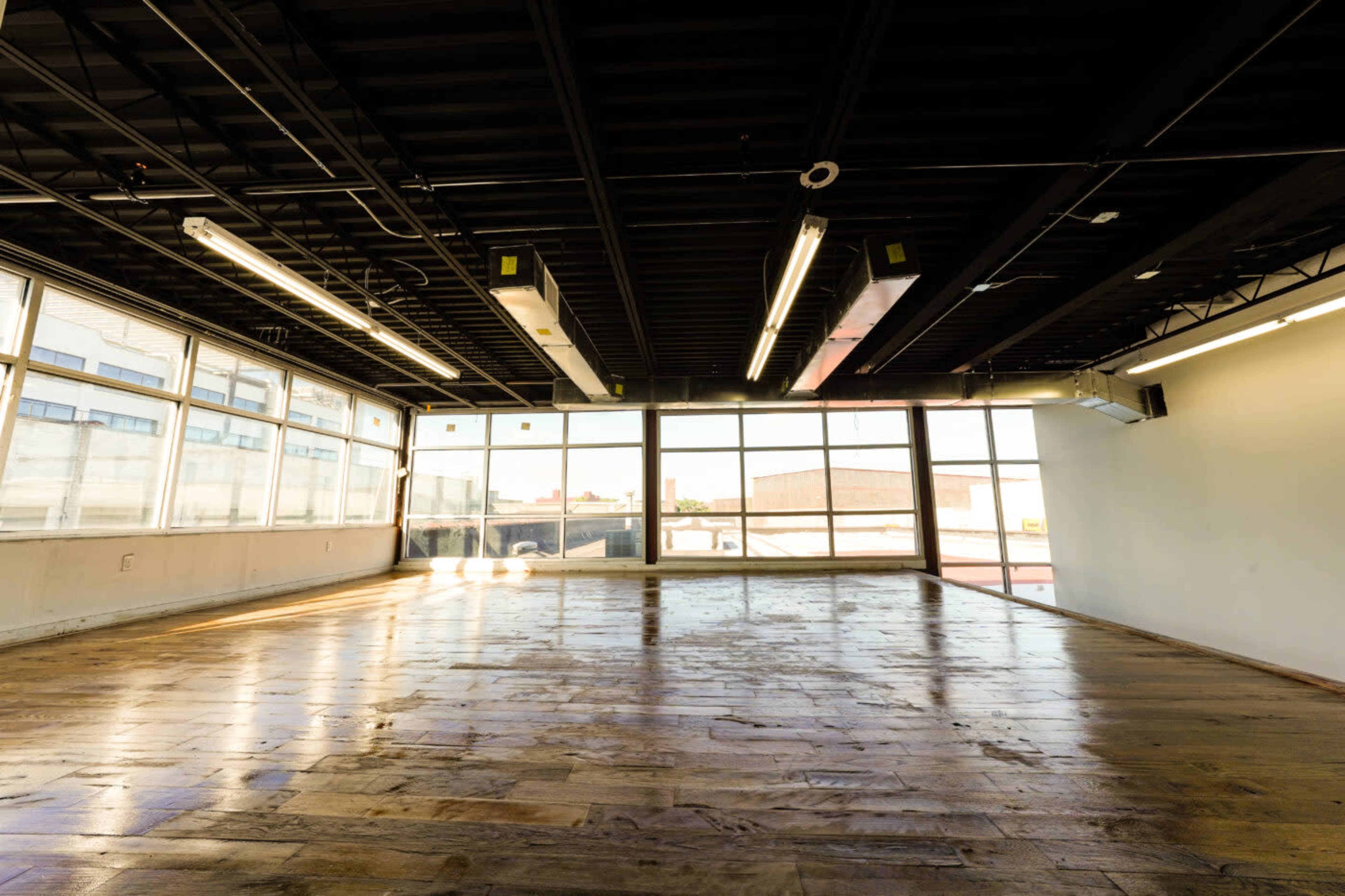 A spacious, empty room with large windows allowing natural light to fill the space and a wooden floor reflecting the light.