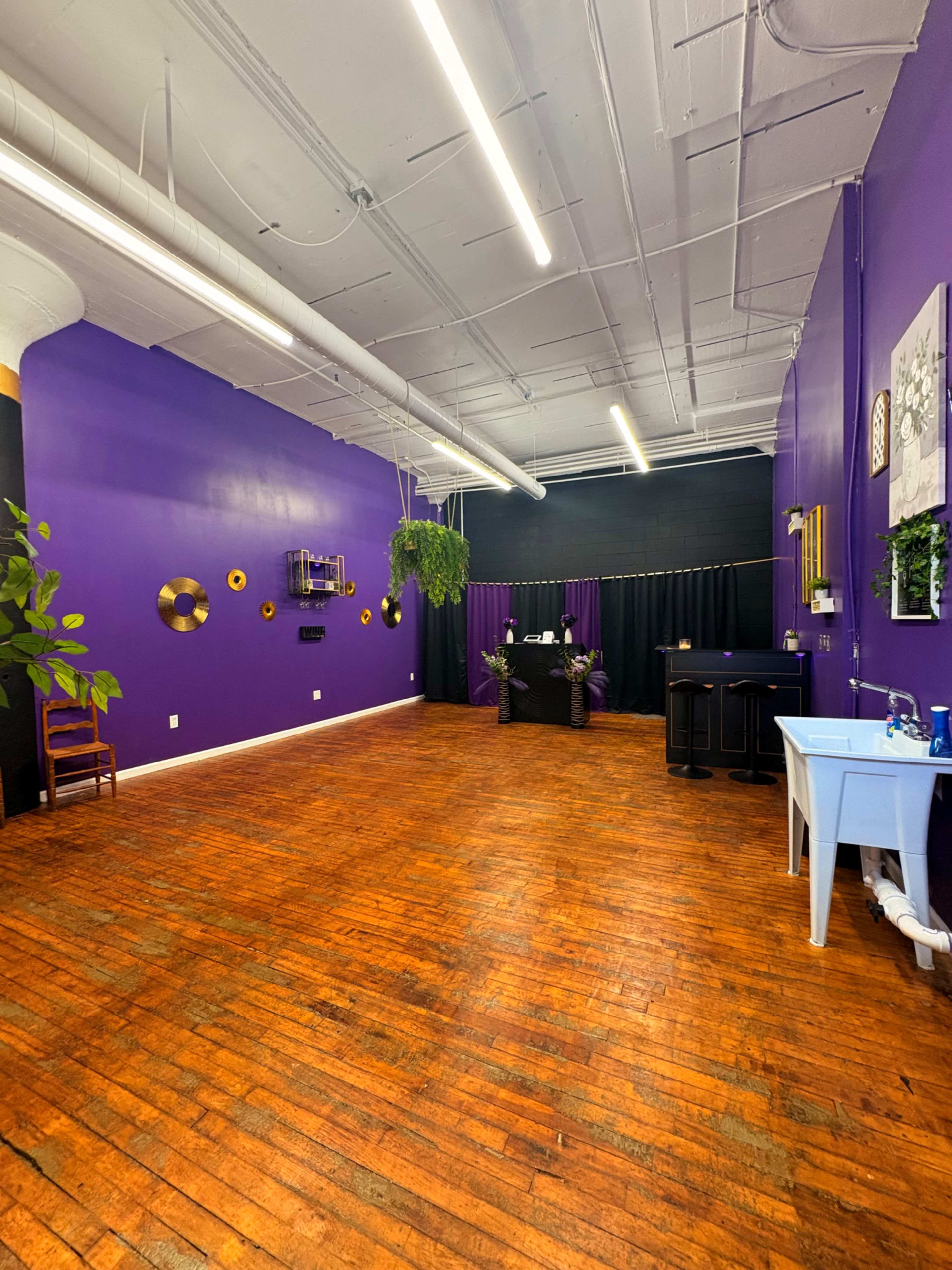 The image shows a spacious room with purple walls, wooden flooring, and various decorations, including plants and art on the walls.