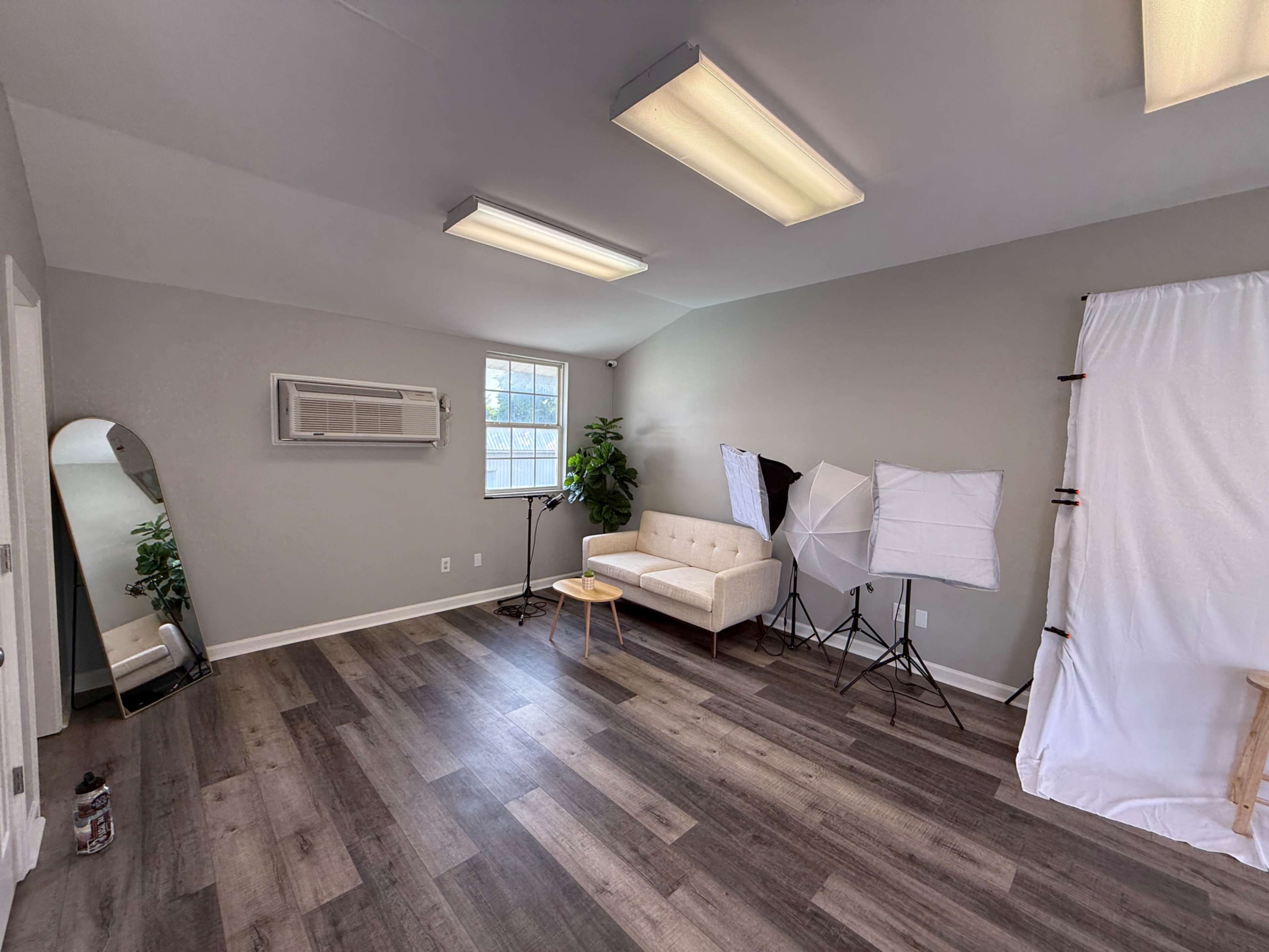 The image shows a spacious, well-lit room featuring a beige couch, an air conditioning unit, a potted plant, and two photography softboxes with a backdrop setup.