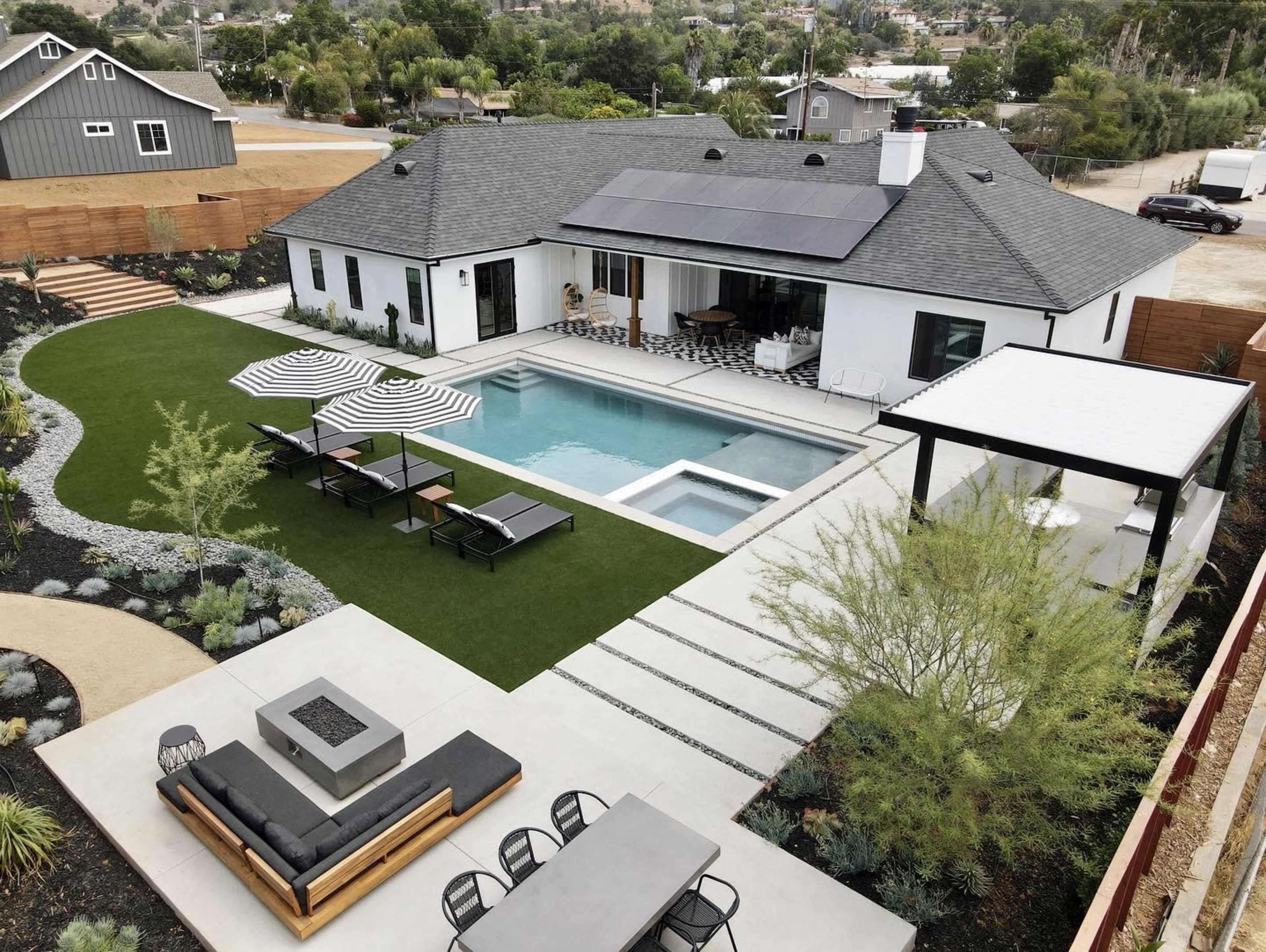 The image showcases a modern backyard featuring a swimming pool, lounge chairs with umbrellas, a fire pit area, and a landscaped garden surrounding a contemporary house with solar panels on the roof.