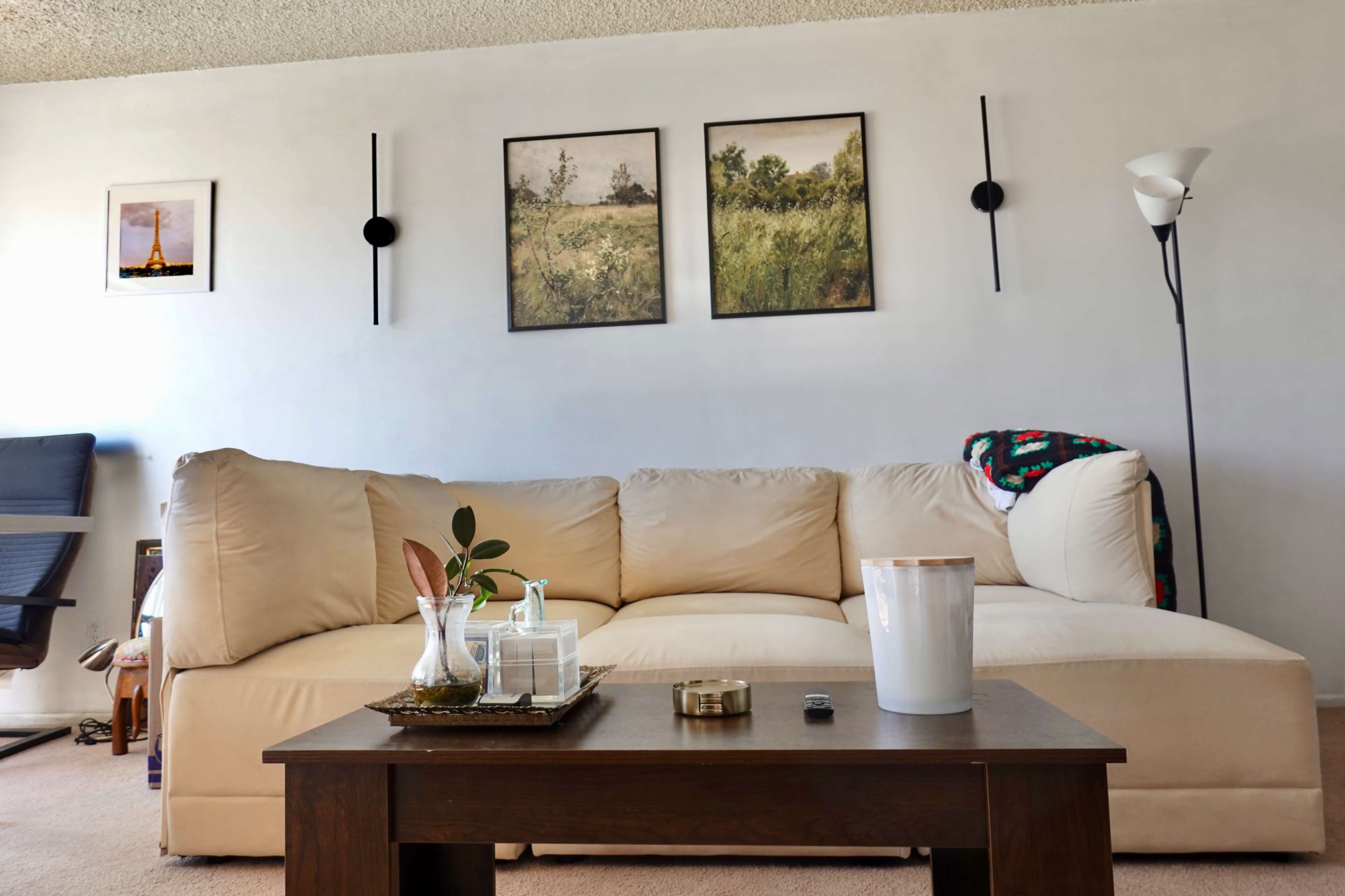 The image shows a light beige sofa against a white wall, with three framed pictures above it and a wooden coffee table in front, featuring decorative items and a lamp nearby.