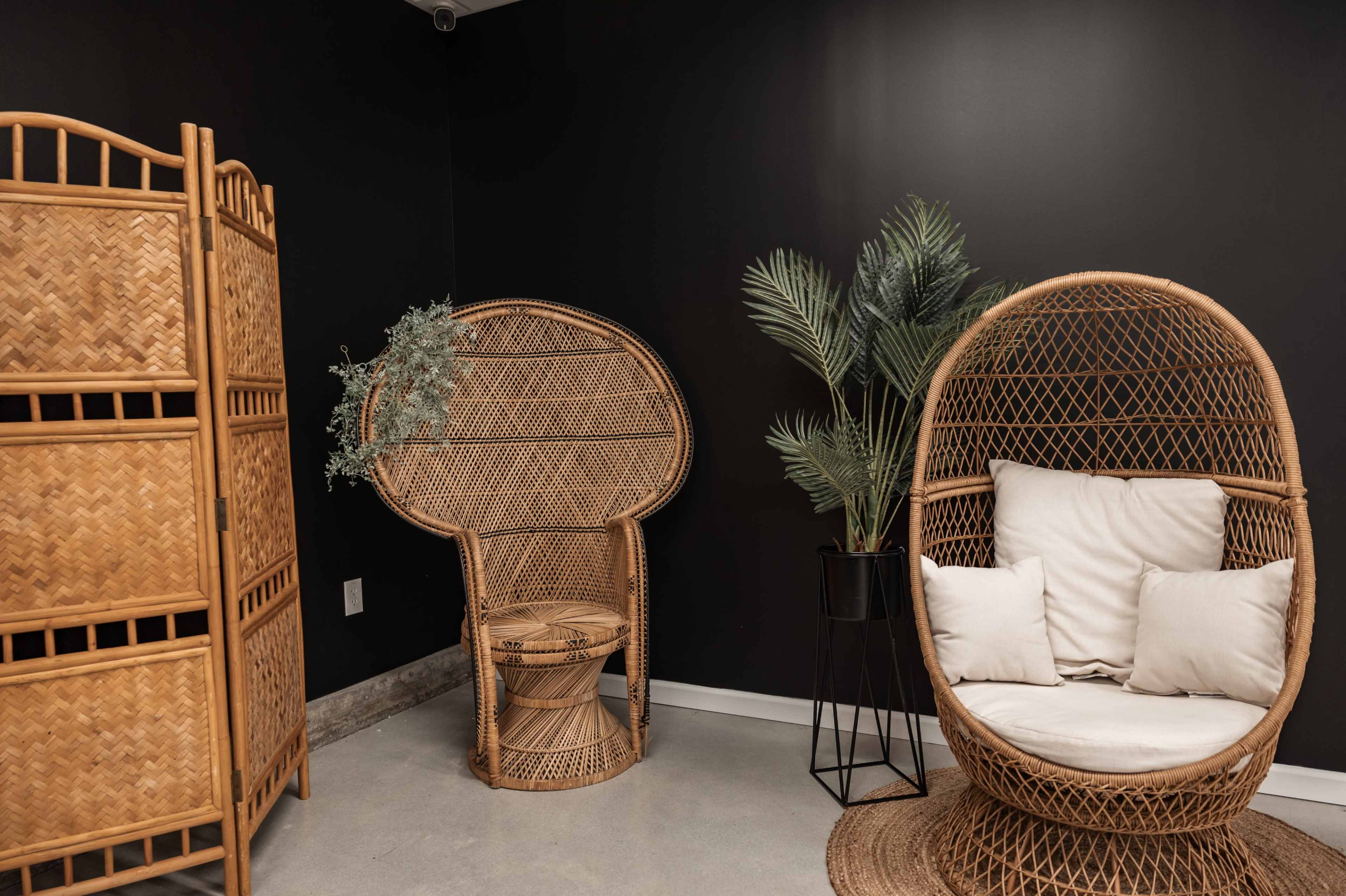 A cozy interior space featuring rattan furniture, including an oversized chair and a circular side table, alongside decorative plants and a folding privacy screen against a black wall.