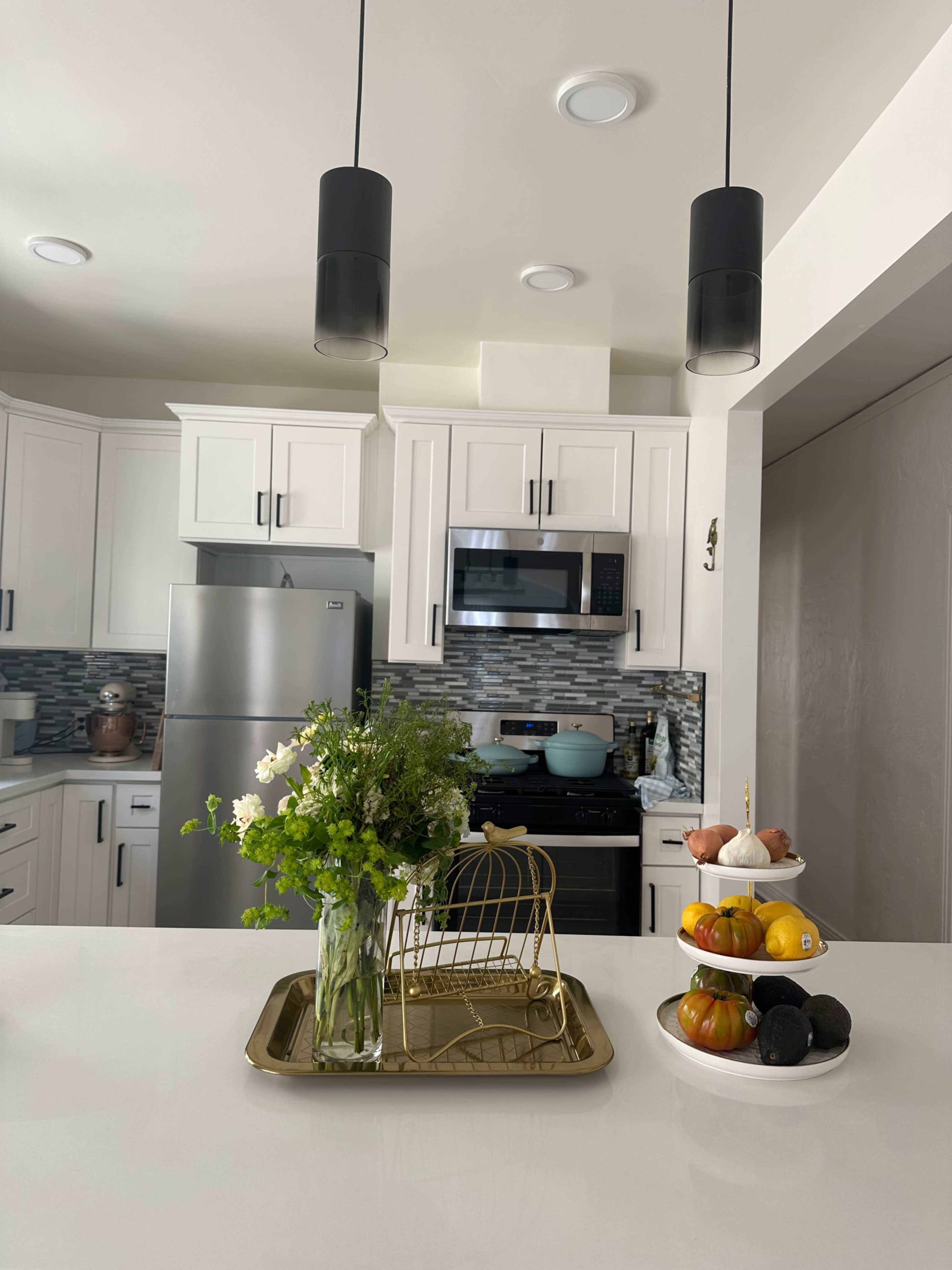 A modern kitchen features sleek white cabinets, stainless steel appliances, and a countertop adorned with a floral arrangement and a bowl of fruit.