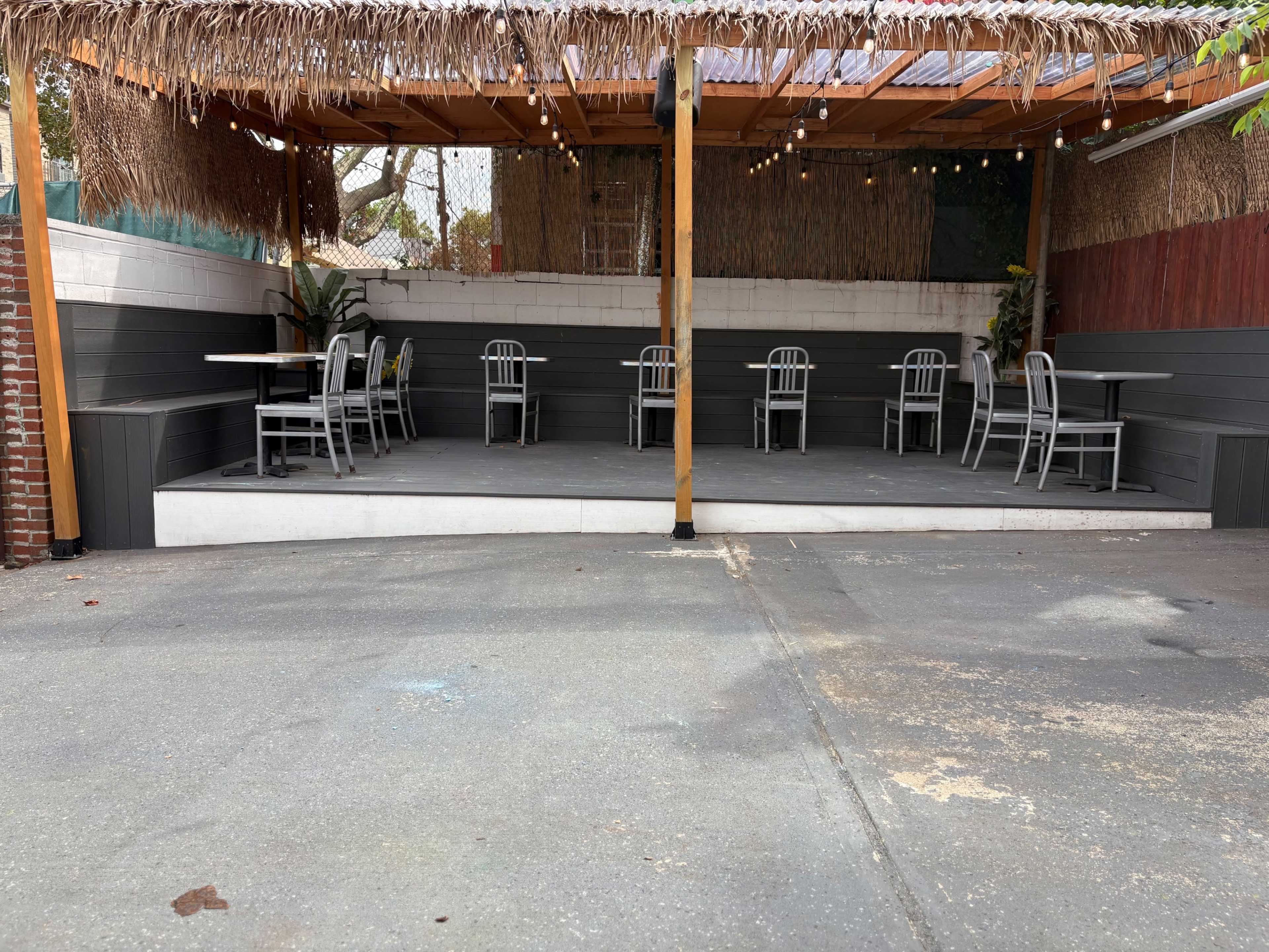The image shows an outdoor seating area of a restaurant with several chairs and tables arranged beneath a thatched roof.