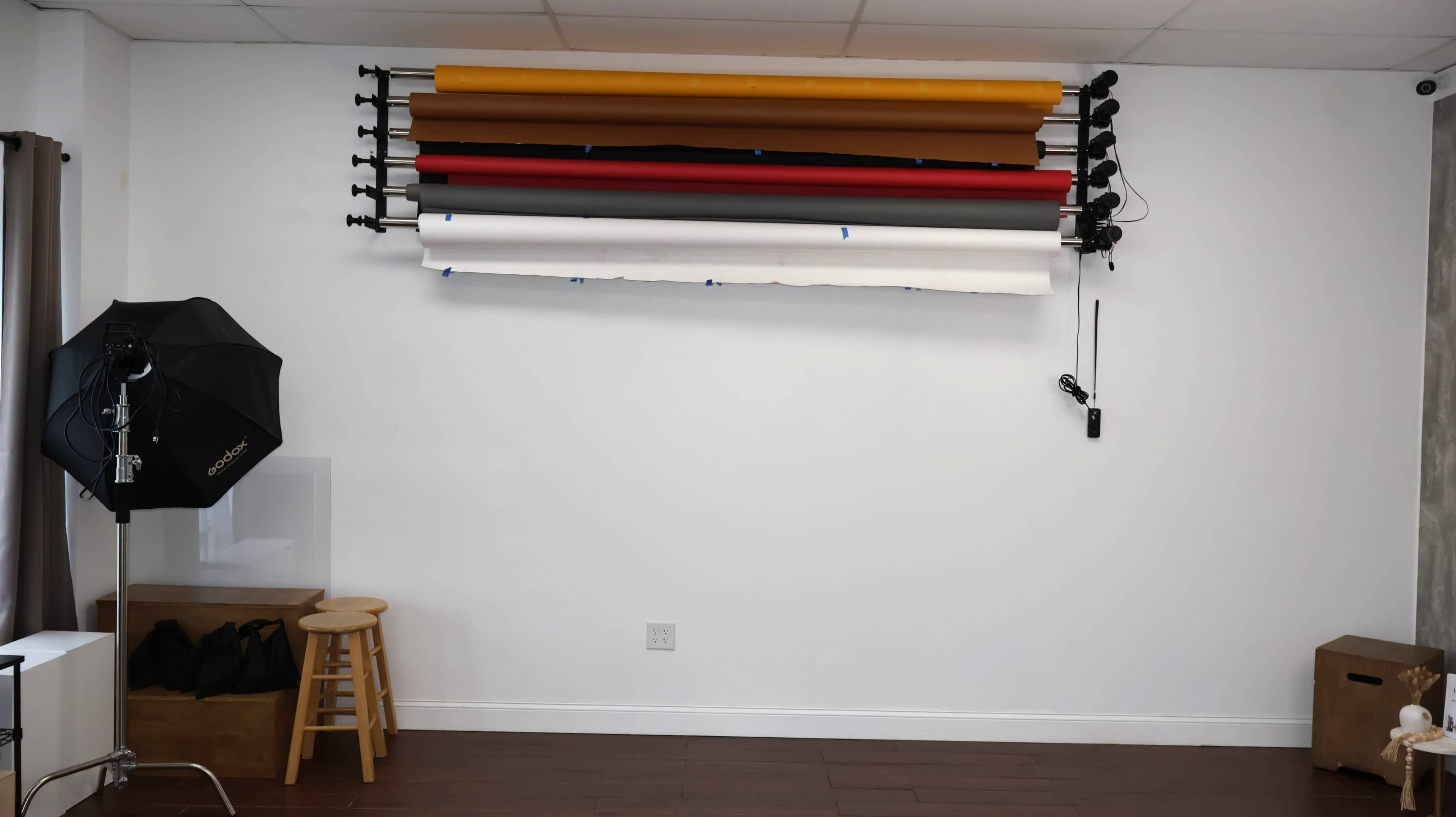 The image shows a studio space with a wall-mounted backdrop system featuring rolls of colored paper, alongside a stool and lighting equipment positioned in the corner.