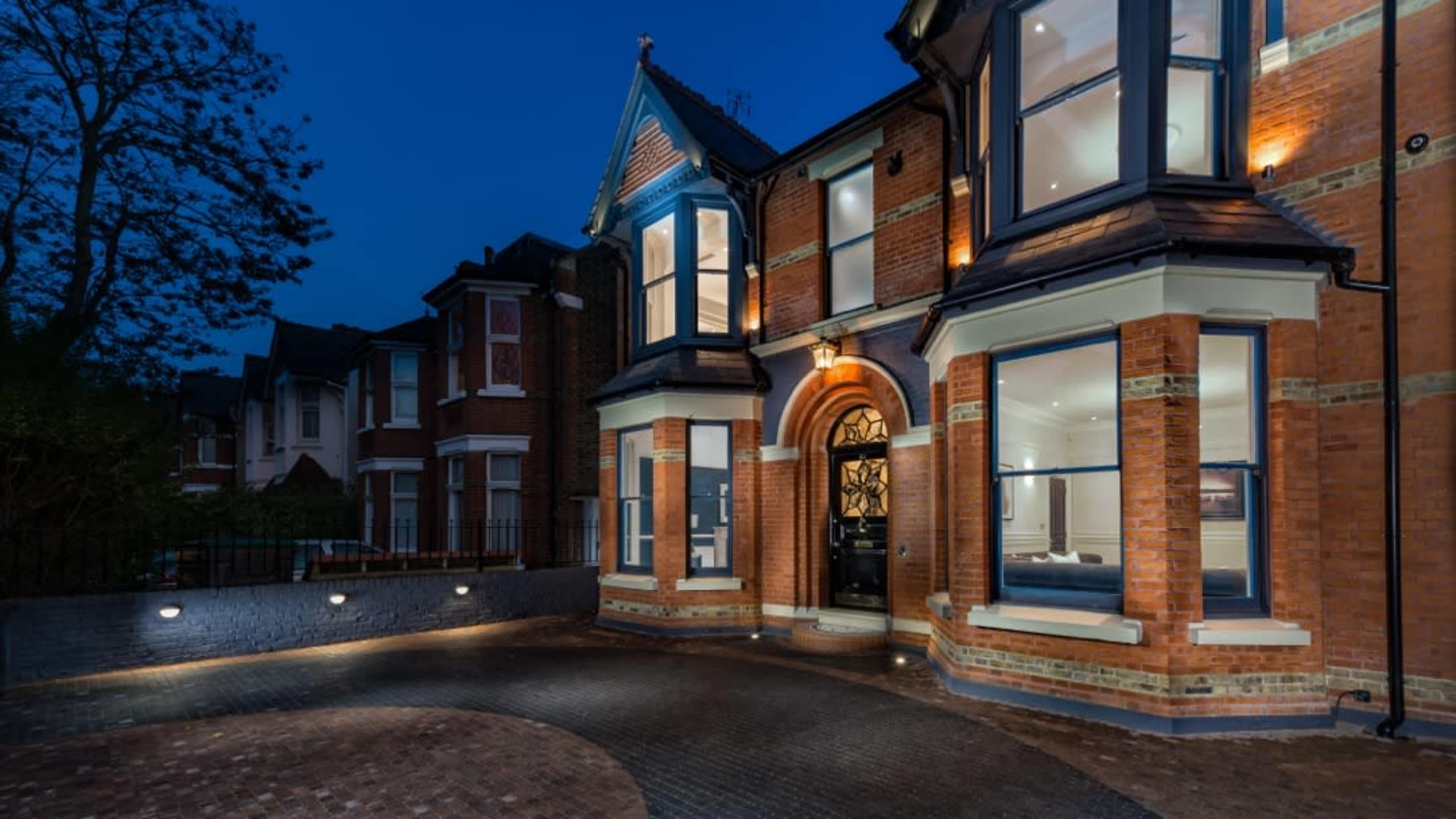 A large, brick-faced house with a prominent entrance and large windows, illuminated against a twilight sky.