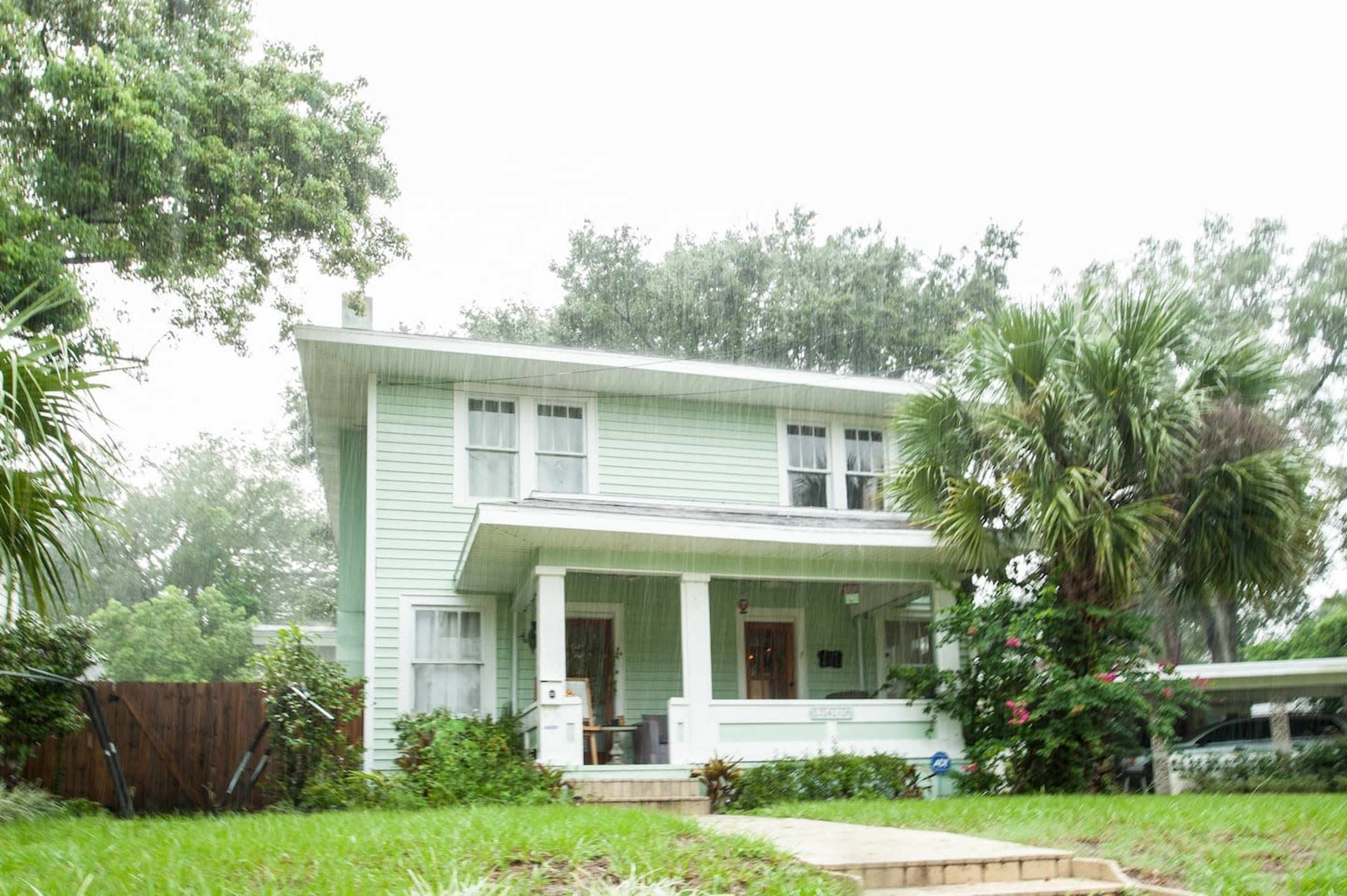 A two-story light green house with white trim and a front porch sits amidst green grass and palm trees in a rainy setting.