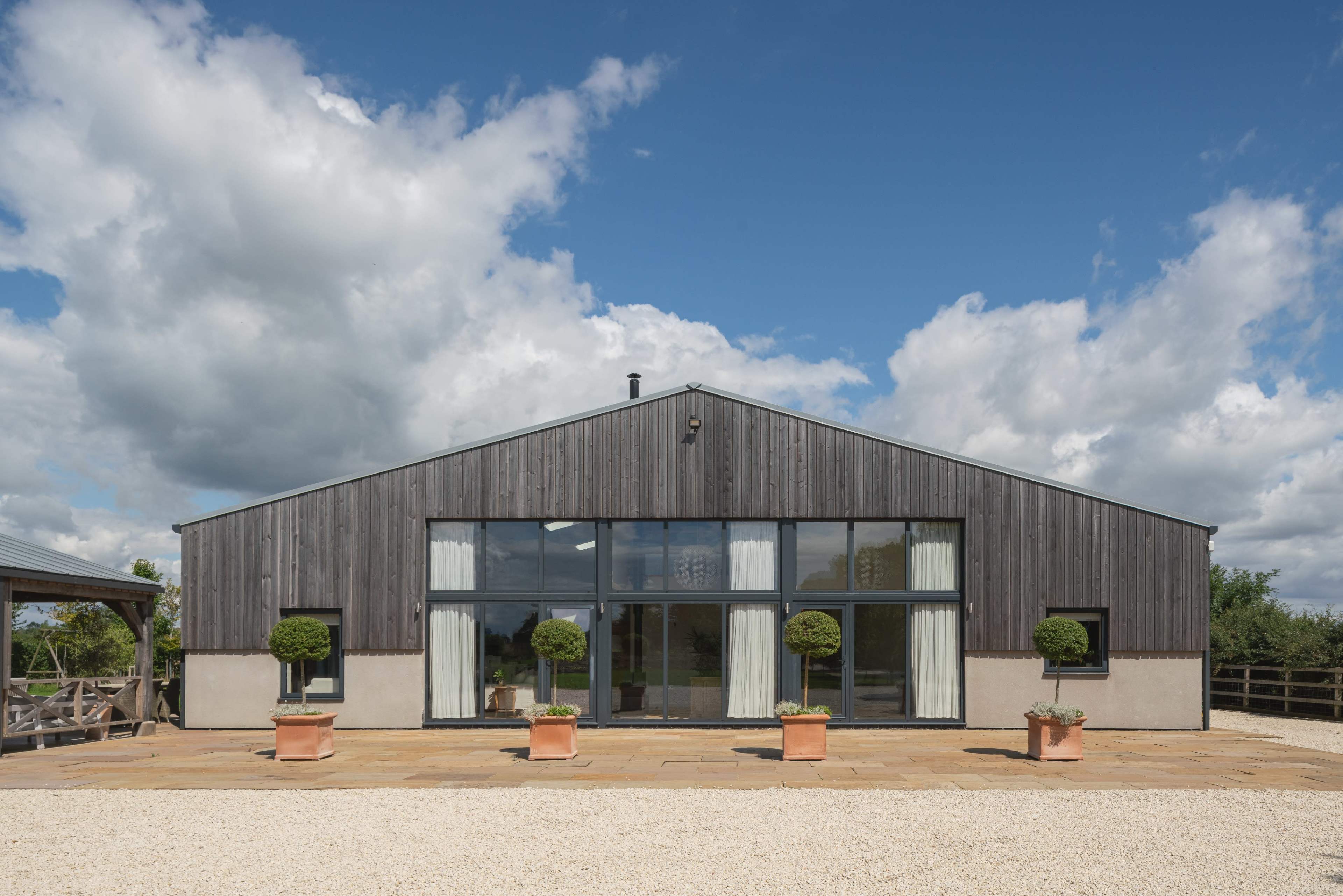 A modern barn-style house with a wooden facade, large glass windows, and potted topiary plants in front, set against a cloudy sky.