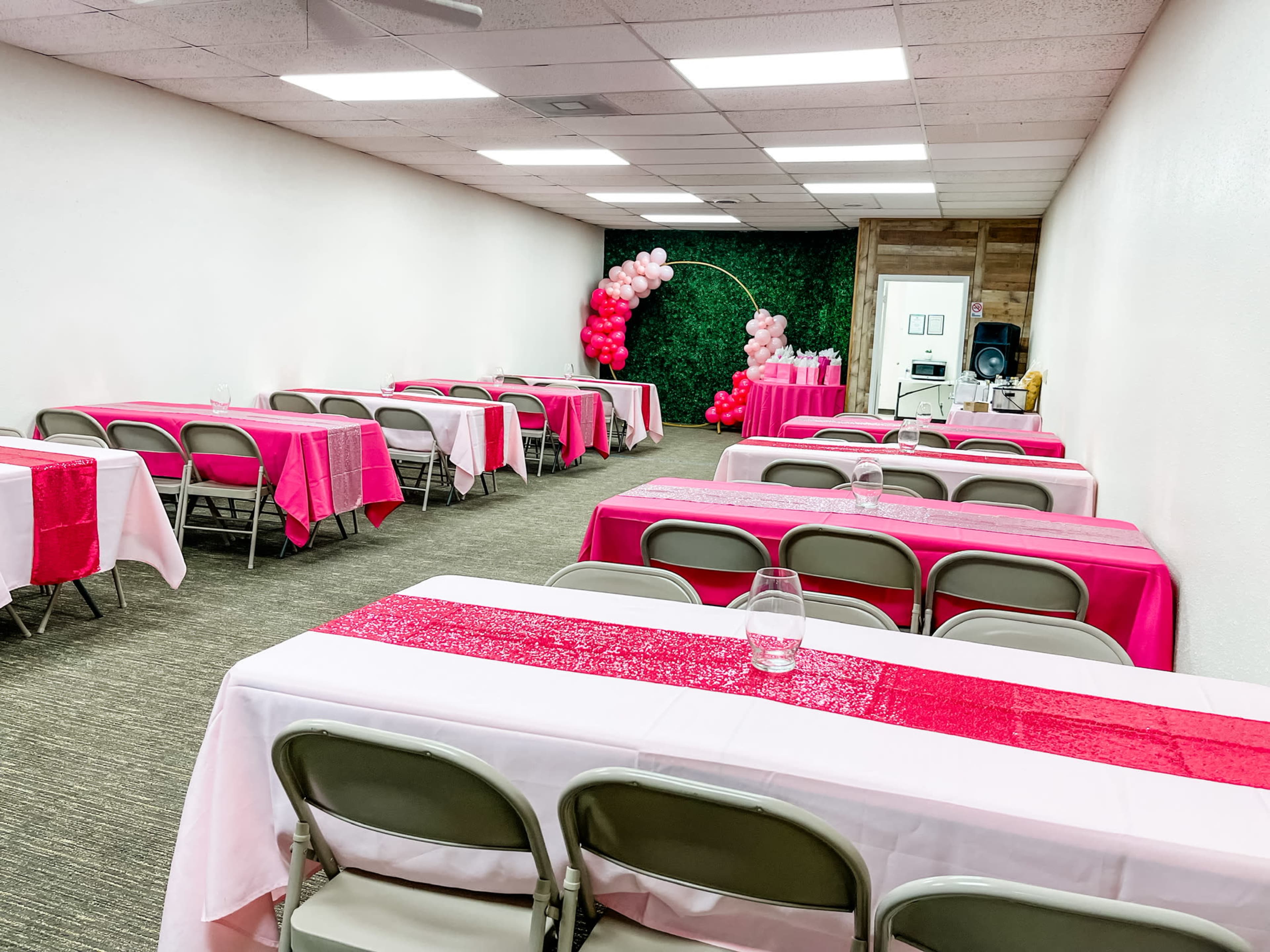 A neatly arranged event space with several tables covered in pink and white tablecloths, facing a decorative green wall and a balloon arrangement in the corner.