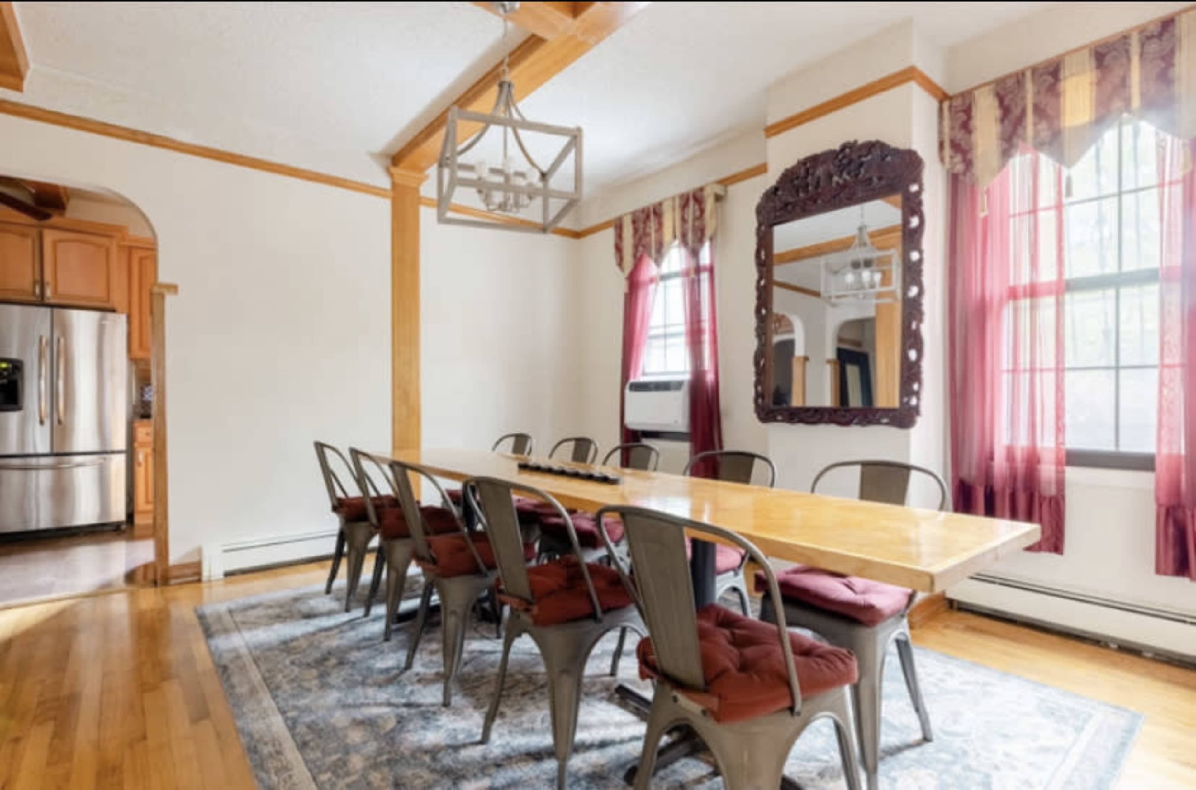 A long wooden dining table is surrounded by metal chairs with cushions, under a chandelier in a room with large windows and patterned curtains.