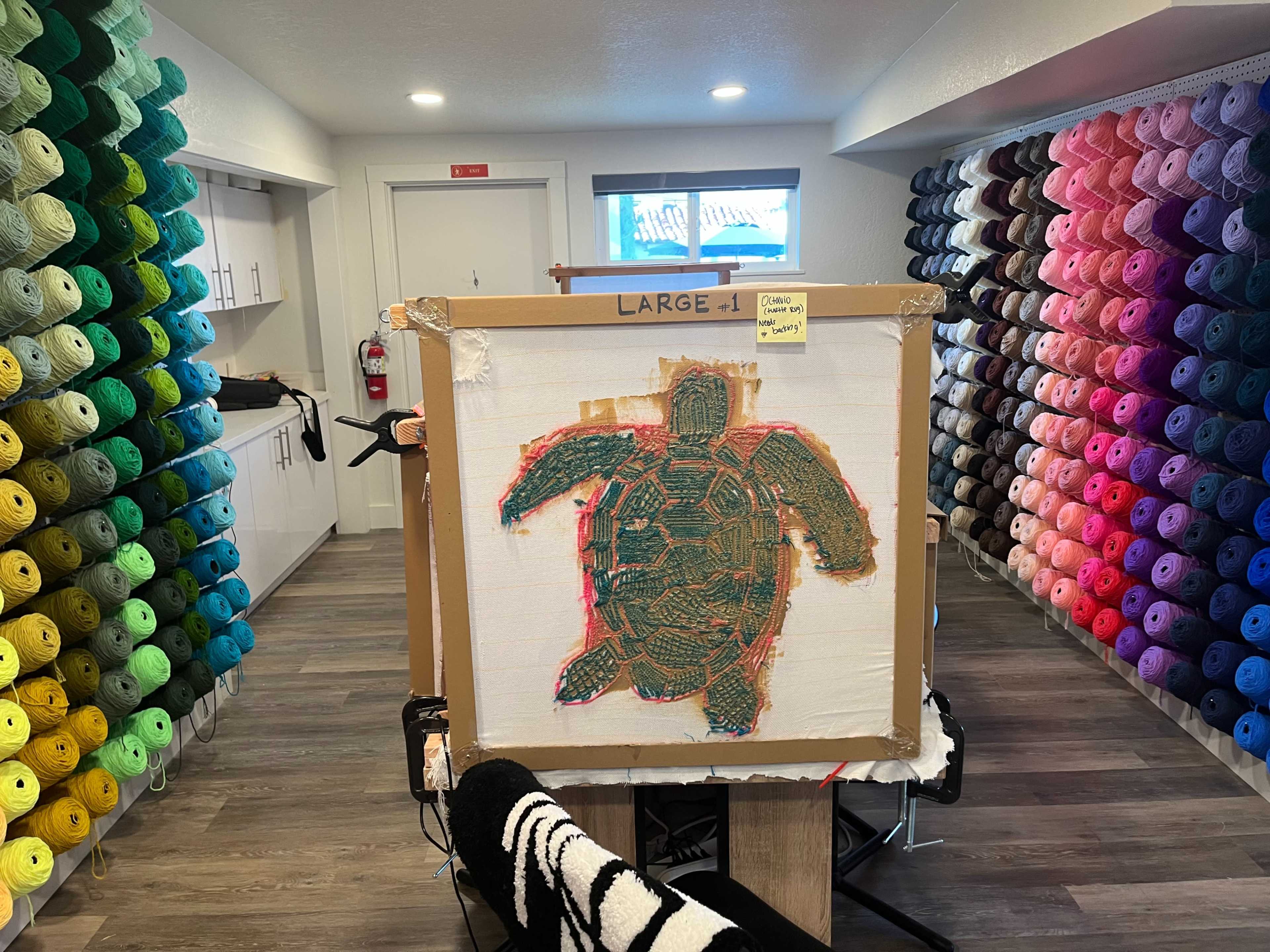 A large canvas depicting a turtle is displayed in front of a wall lined with colorful rolls of yarn.