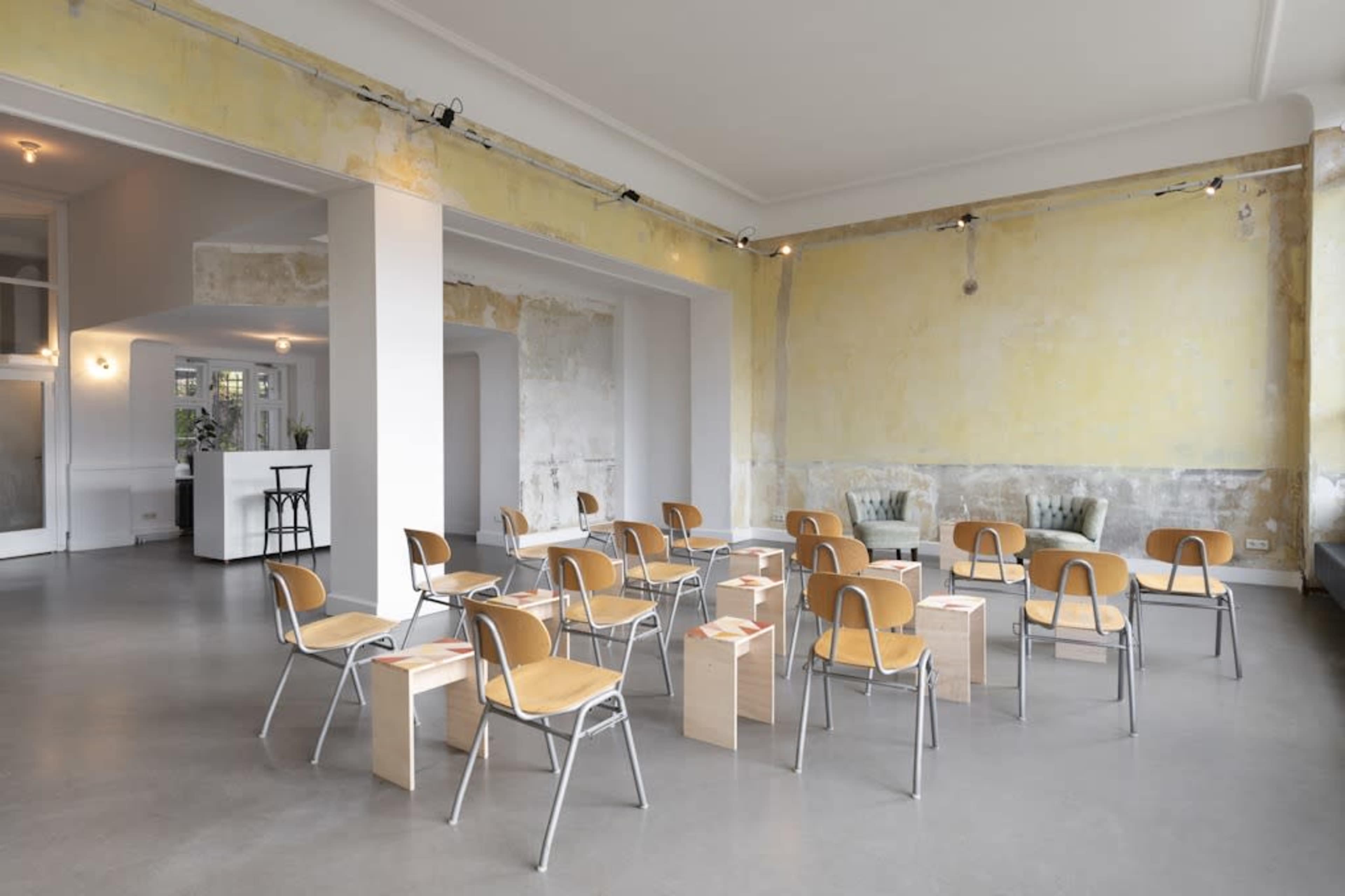 A spacious, minimalist room with several metal and wooden chairs arranged around wooden stools in front of a pale yellow wall.