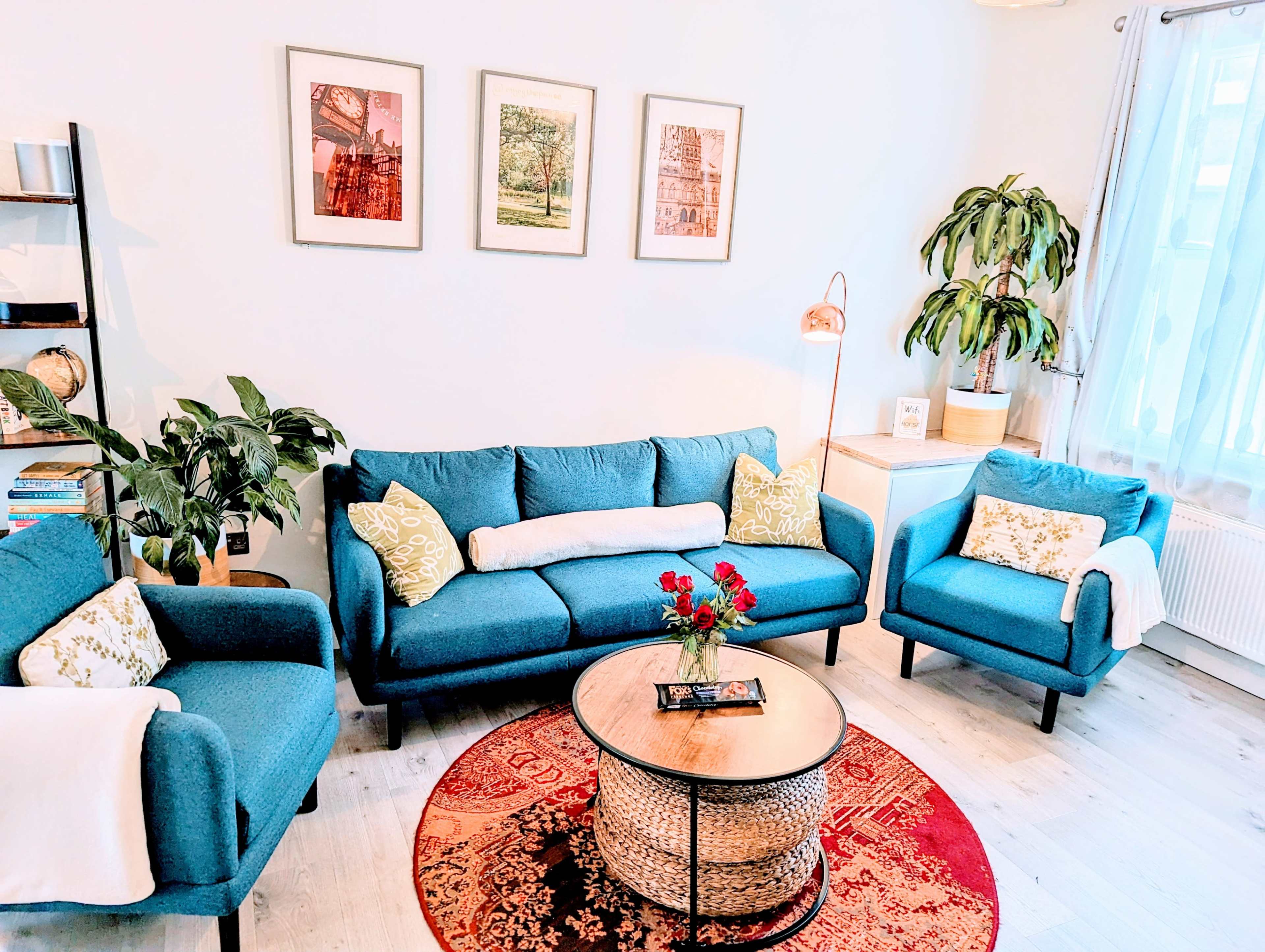 A cozy living room features a blue sofa and armchairs, a round coffee table, a decorative rug, and framed artwork on the walls.