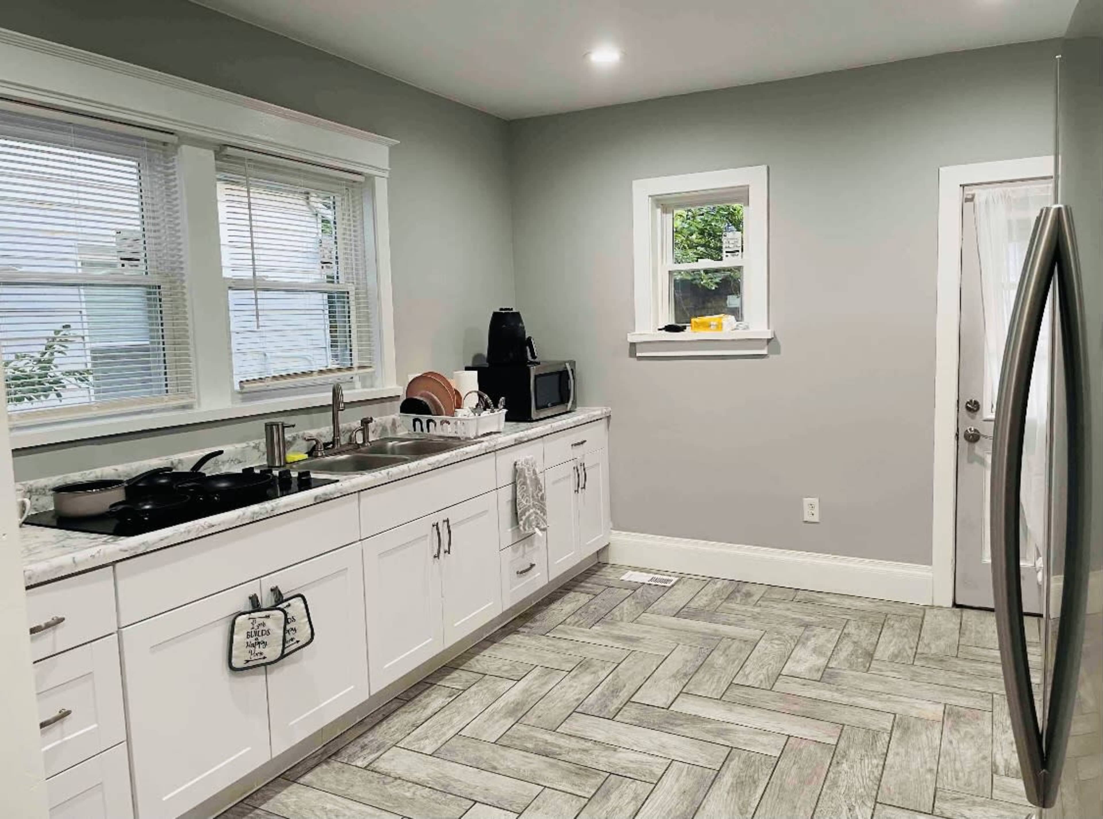 The kitchen features white cabinetry, a large stainless steel refrigerator, and a light gray color scheme with a herringbone patterned floor.