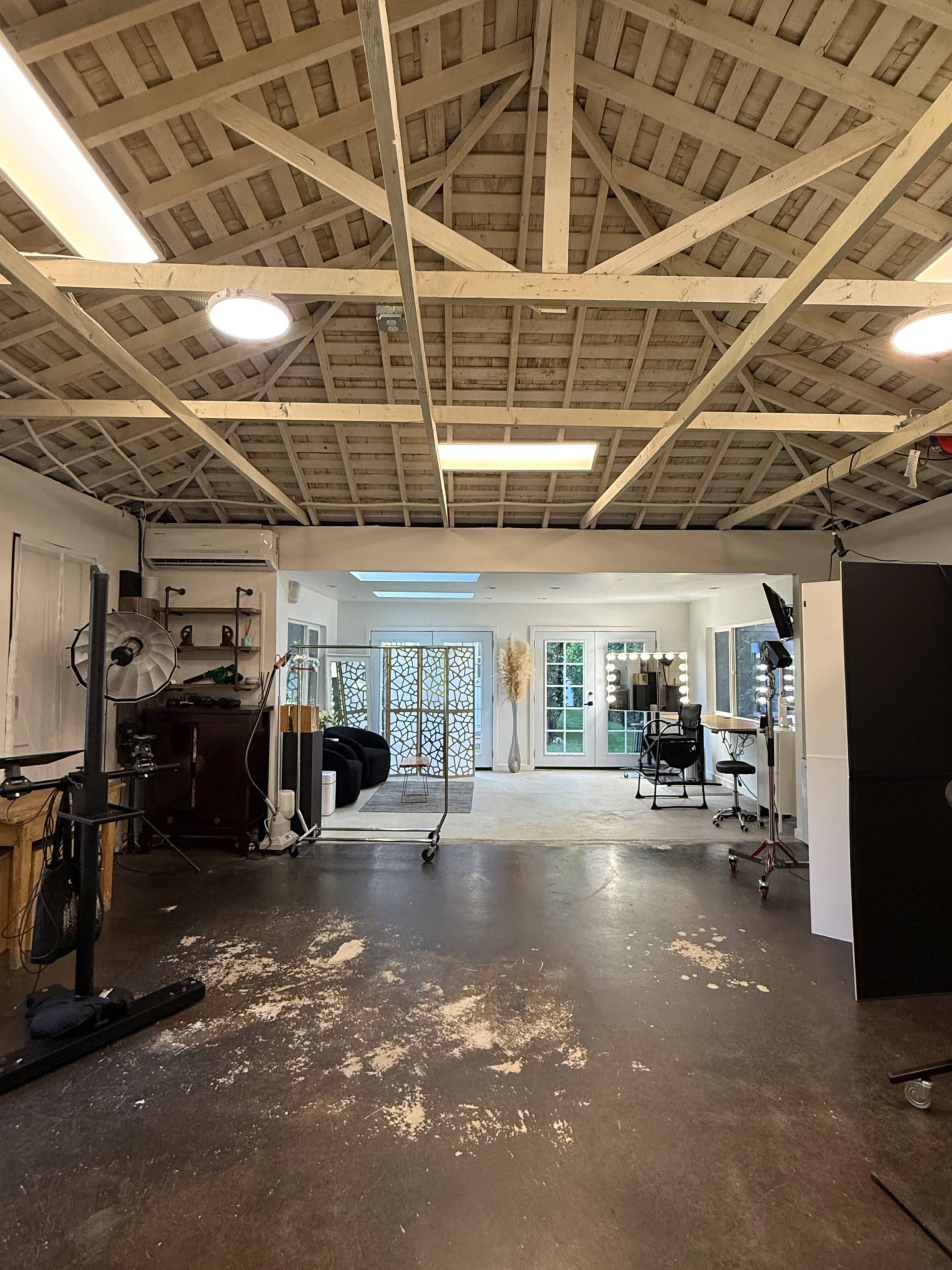 The image shows a spacious studio with a wooden ceiling, polished concrete floor, and various workstations including a makeup station and a seating area.