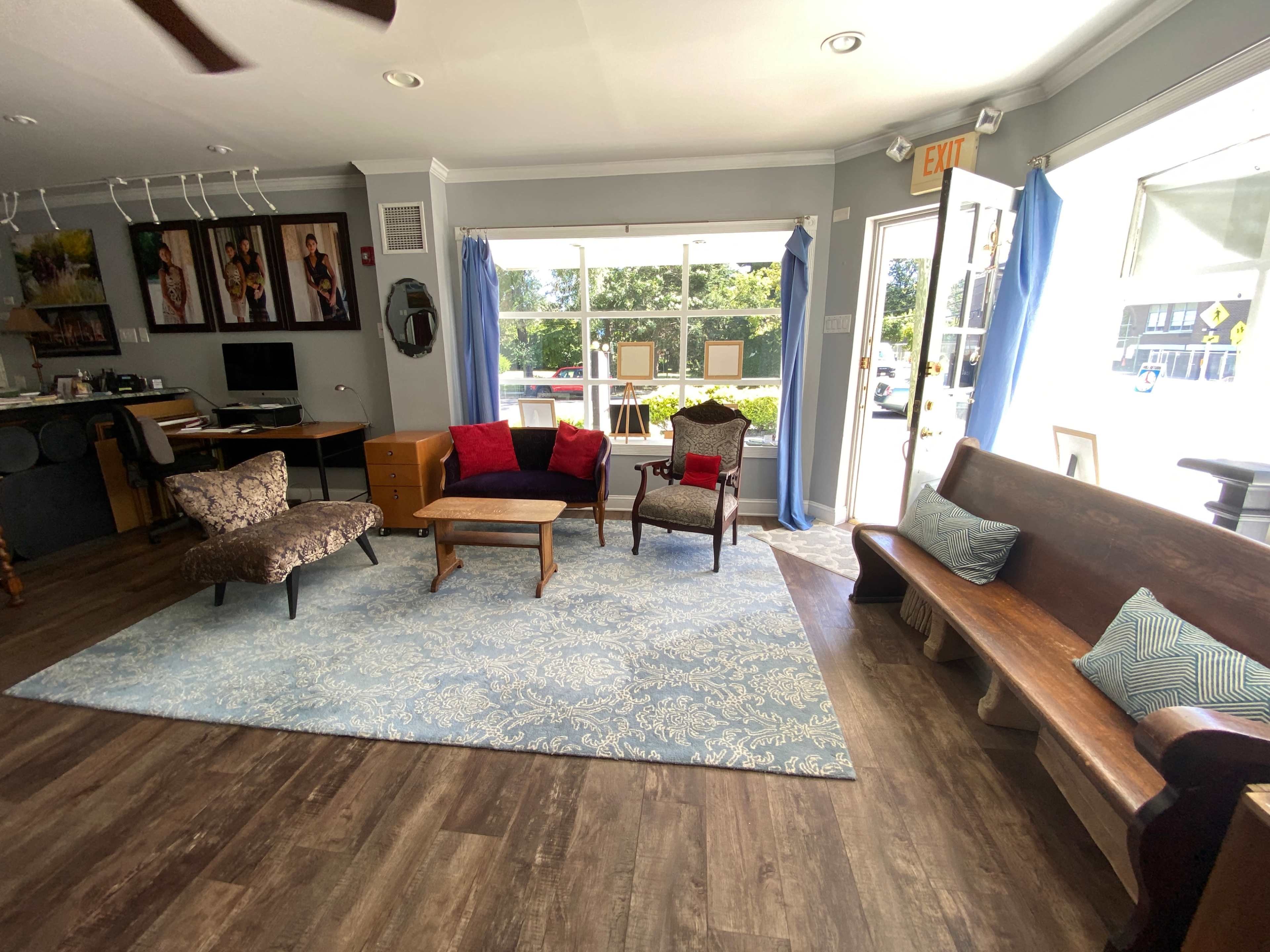 The image shows a spacious room with wooden flooring, featuring various seating options, a decorative rug, and large windows that allow natural light to enter.