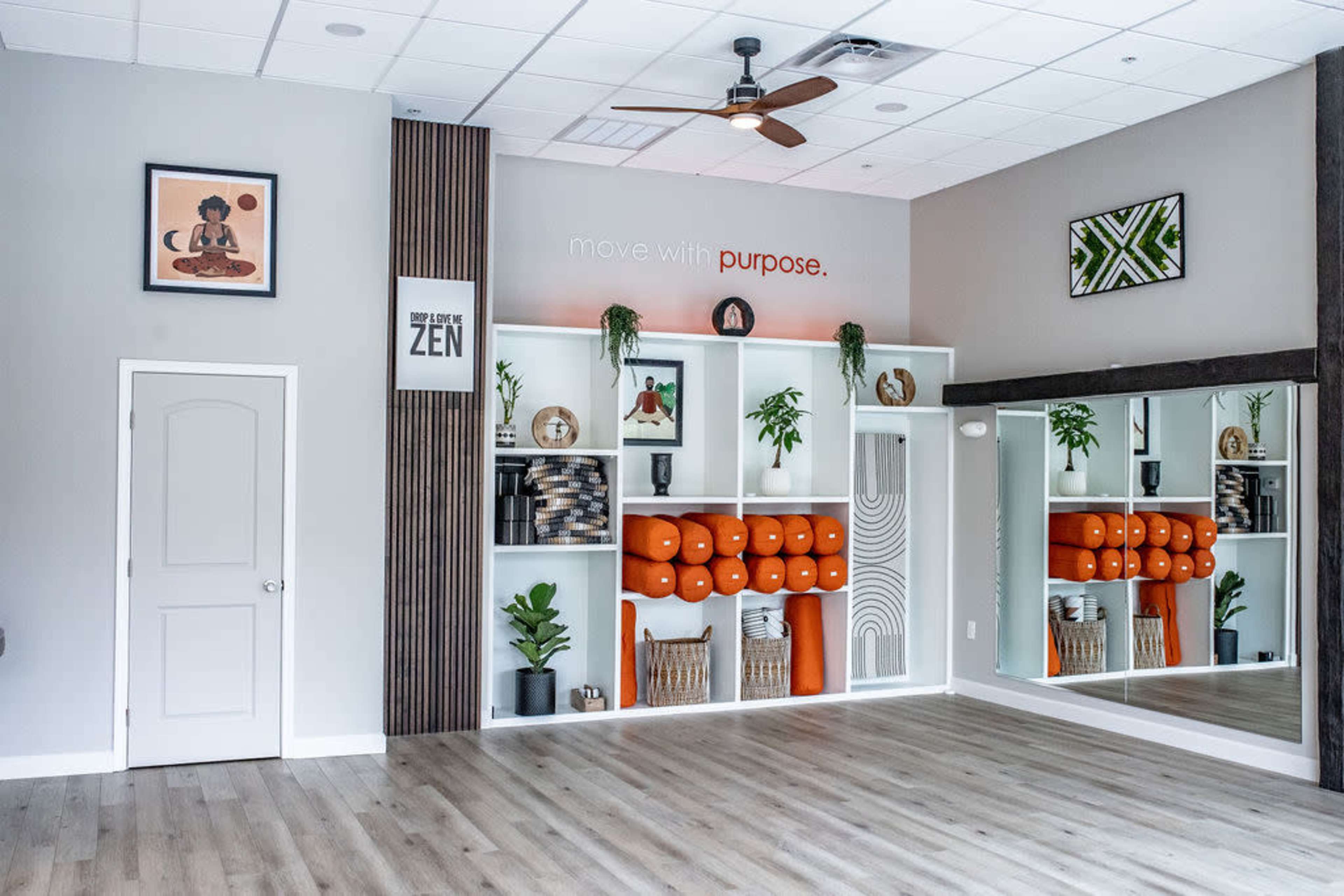A fitness studio features a wall of shelves displaying exercise equipment and greenery, with motivational artwork and a mirrored wall.