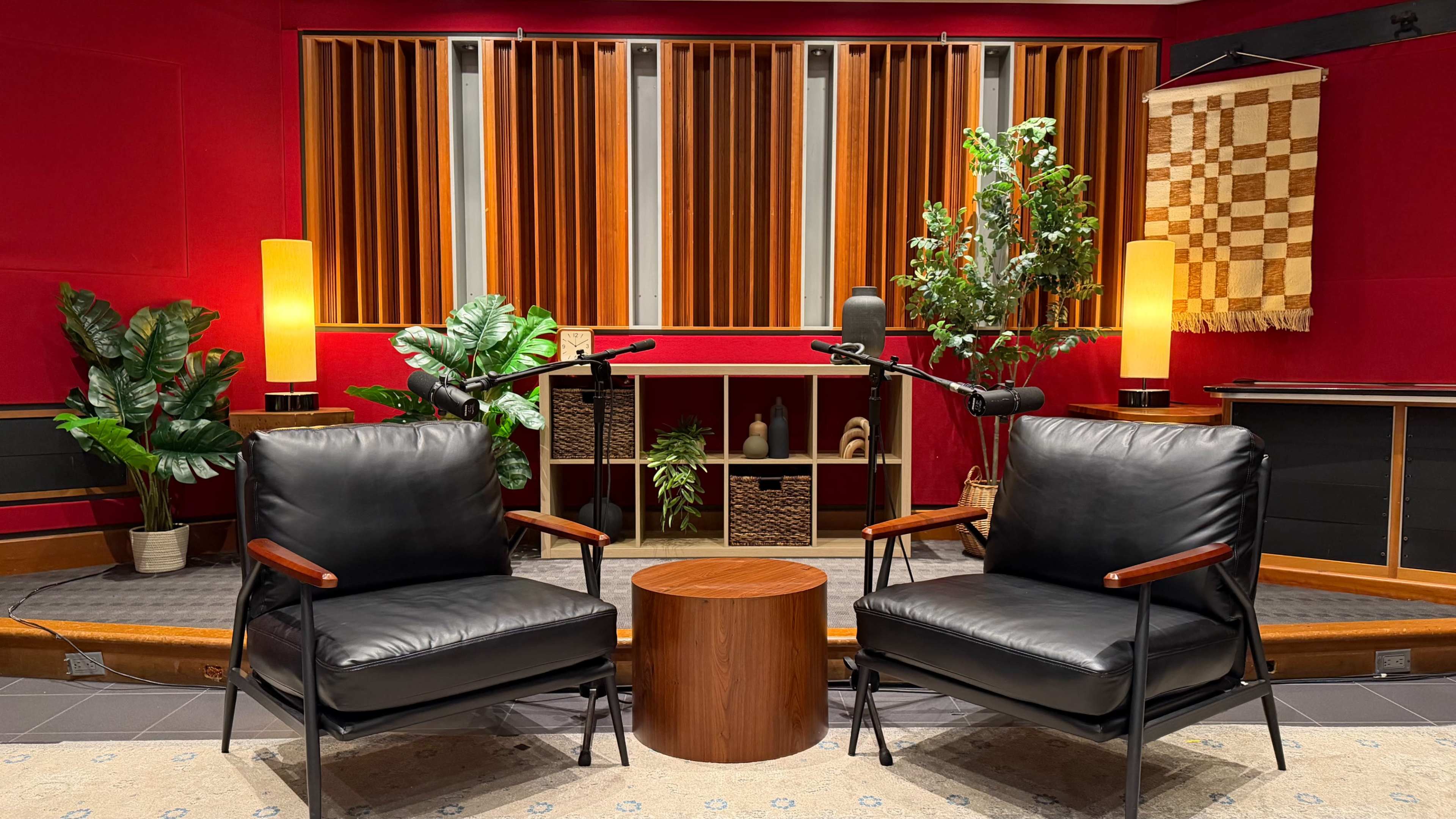 The image shows a cozy audio recording studio with two black chairs facing a wooden table, surrounded by plants, wooden sound panels, and ambient lighting.