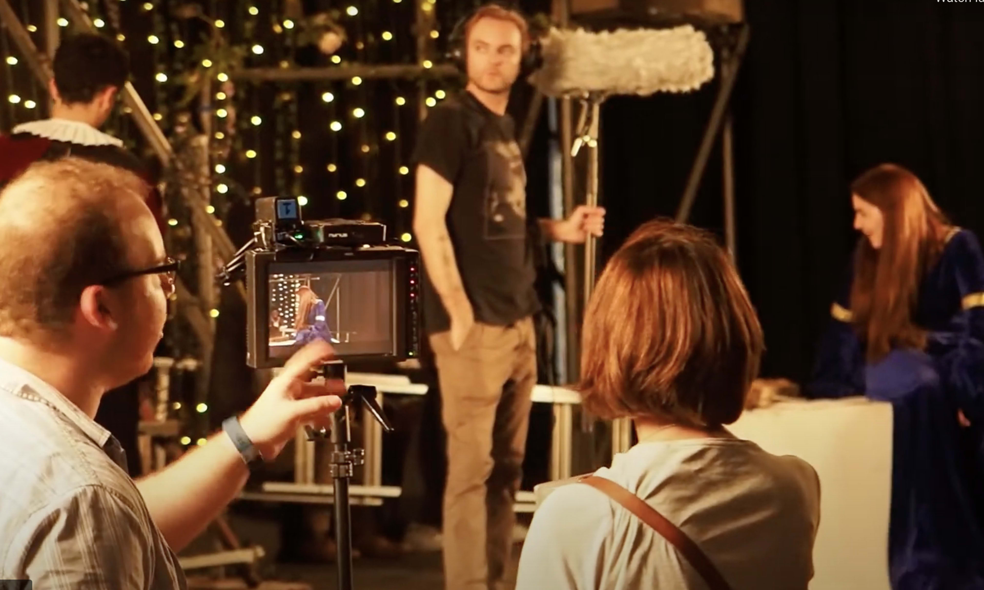 A film crew sets up equipment in a studio with decorative lights while preparing a scene featuring actors in costumes.