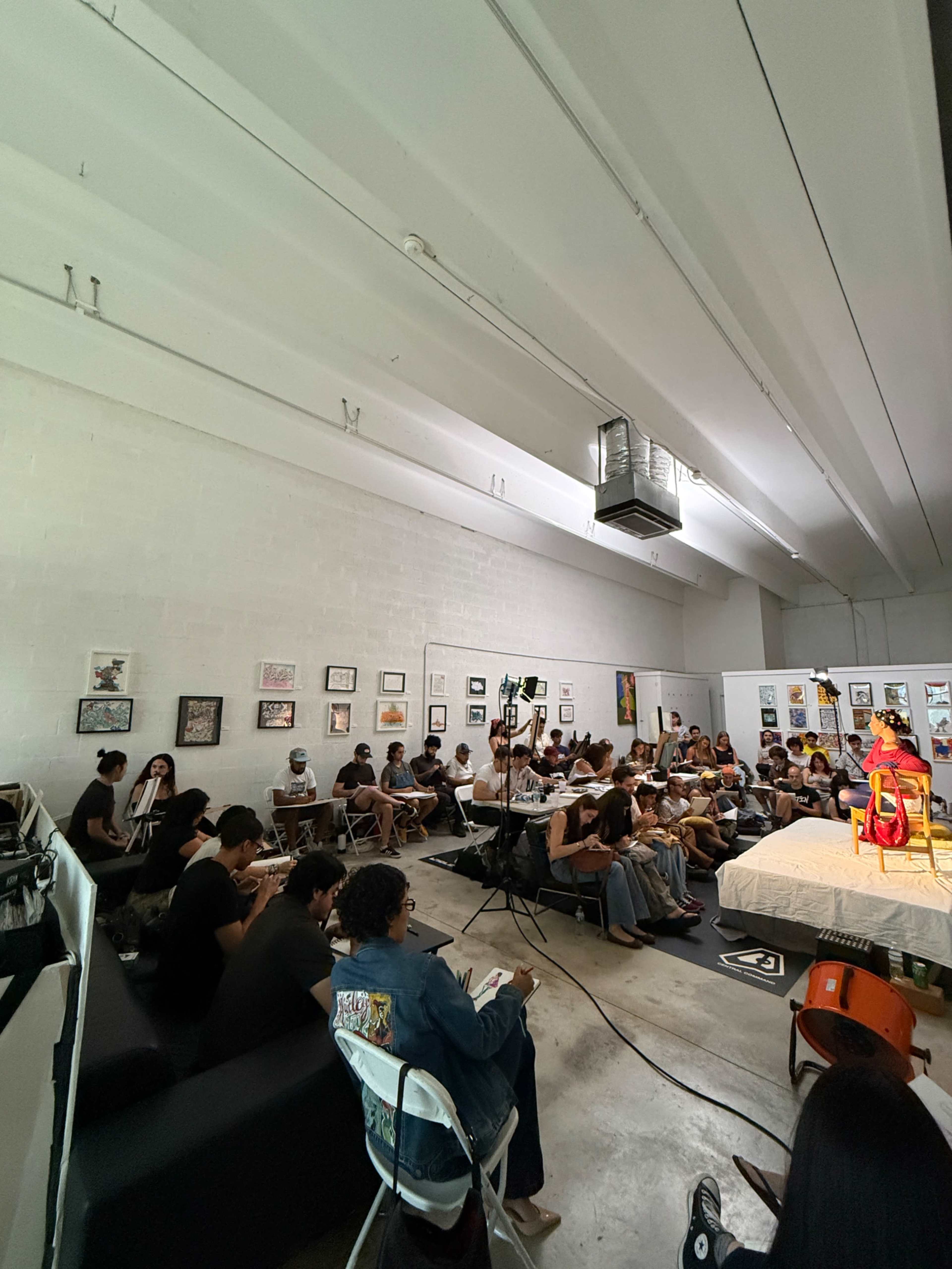 A diverse group of people sits attentively in an art space, facing a speaker on a stage surrounded by various artworks on the walls.