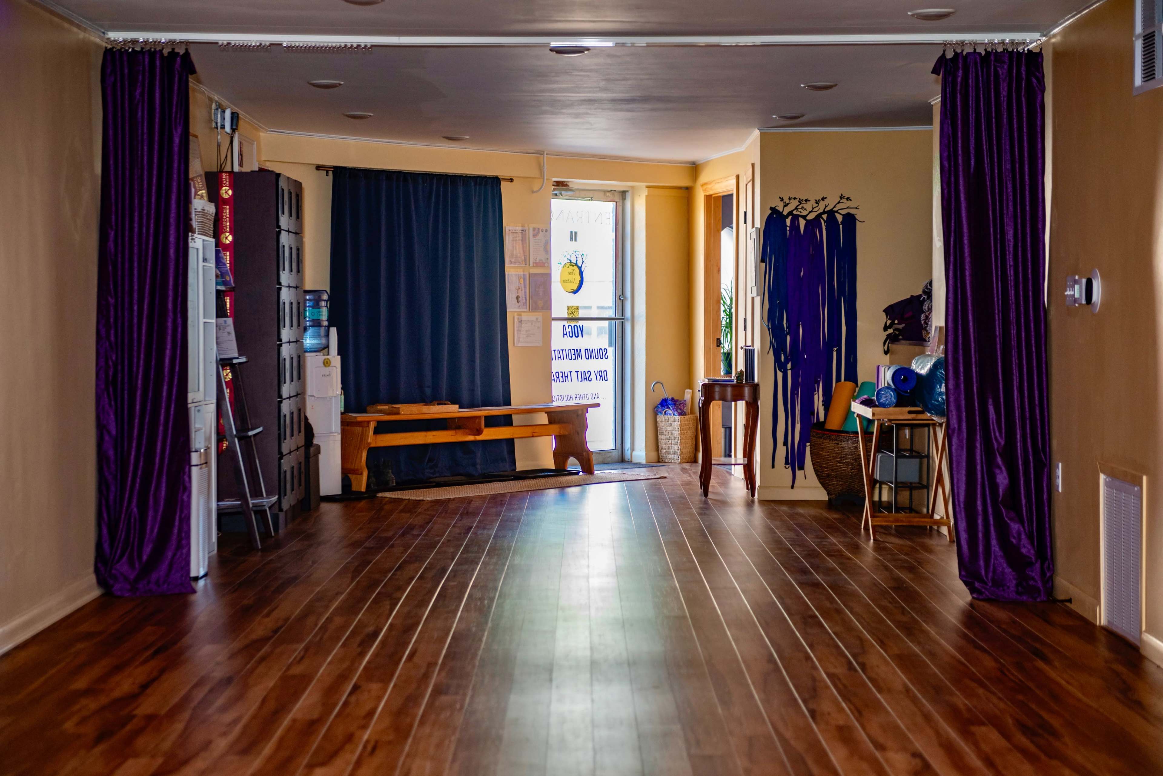 The image shows a spacious, well-lit room with wooden flooring, a large window at one end, and purple curtains along the sides.