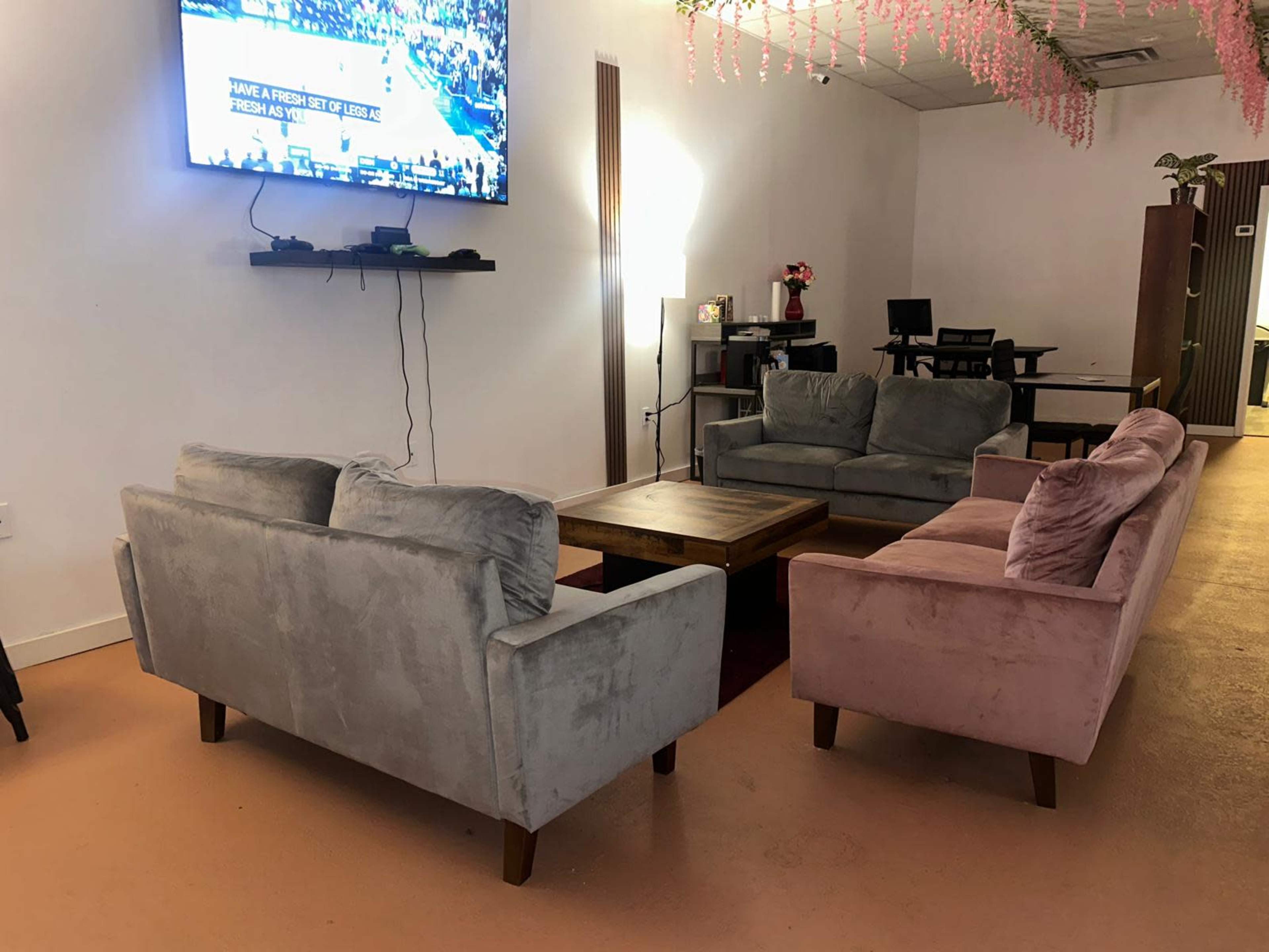 A cozy lounge area features two sofas—one gray and one pink—facing a wooden coffee table, with a TV mounted on the wall and a desk in the background.