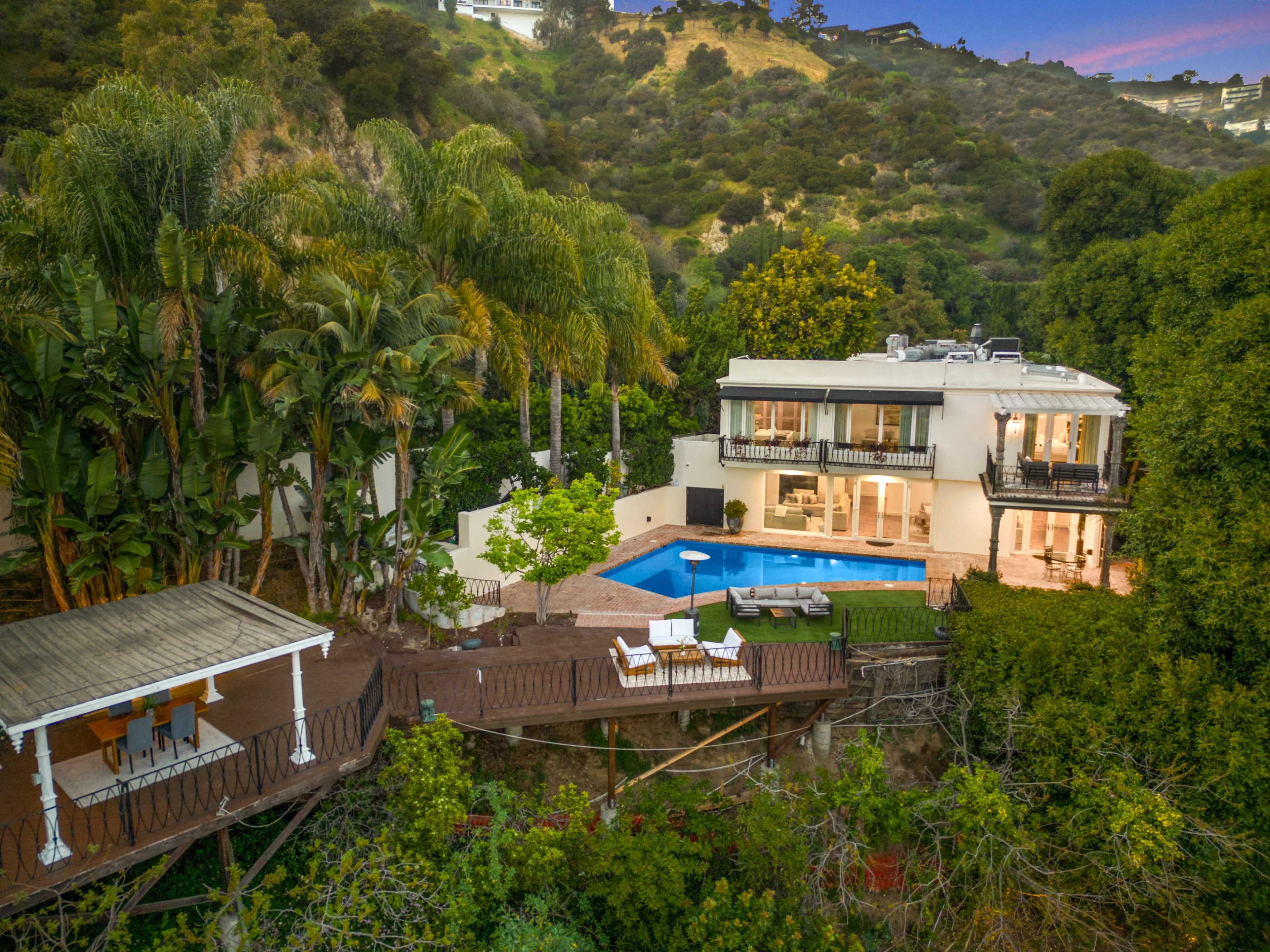 The image shows a multi-level house with a swimming pool and patio area surrounded by lush greenery and trees.