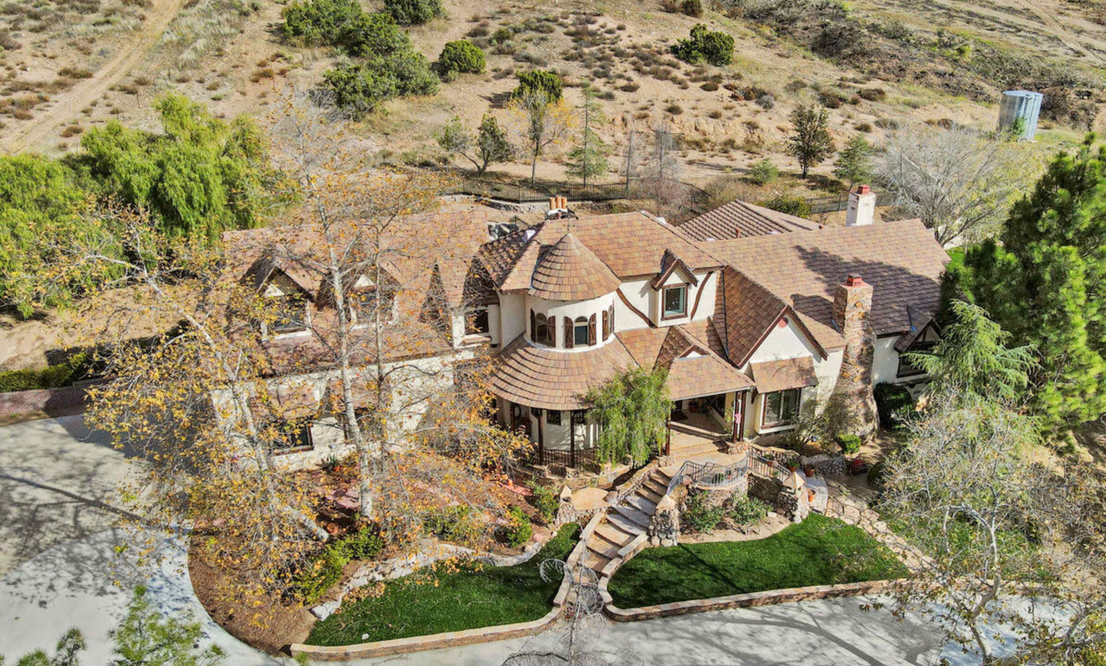 A large, upscale home surrounded by trees and a landscaped yard, with winding pathways and a hilly backdrop.