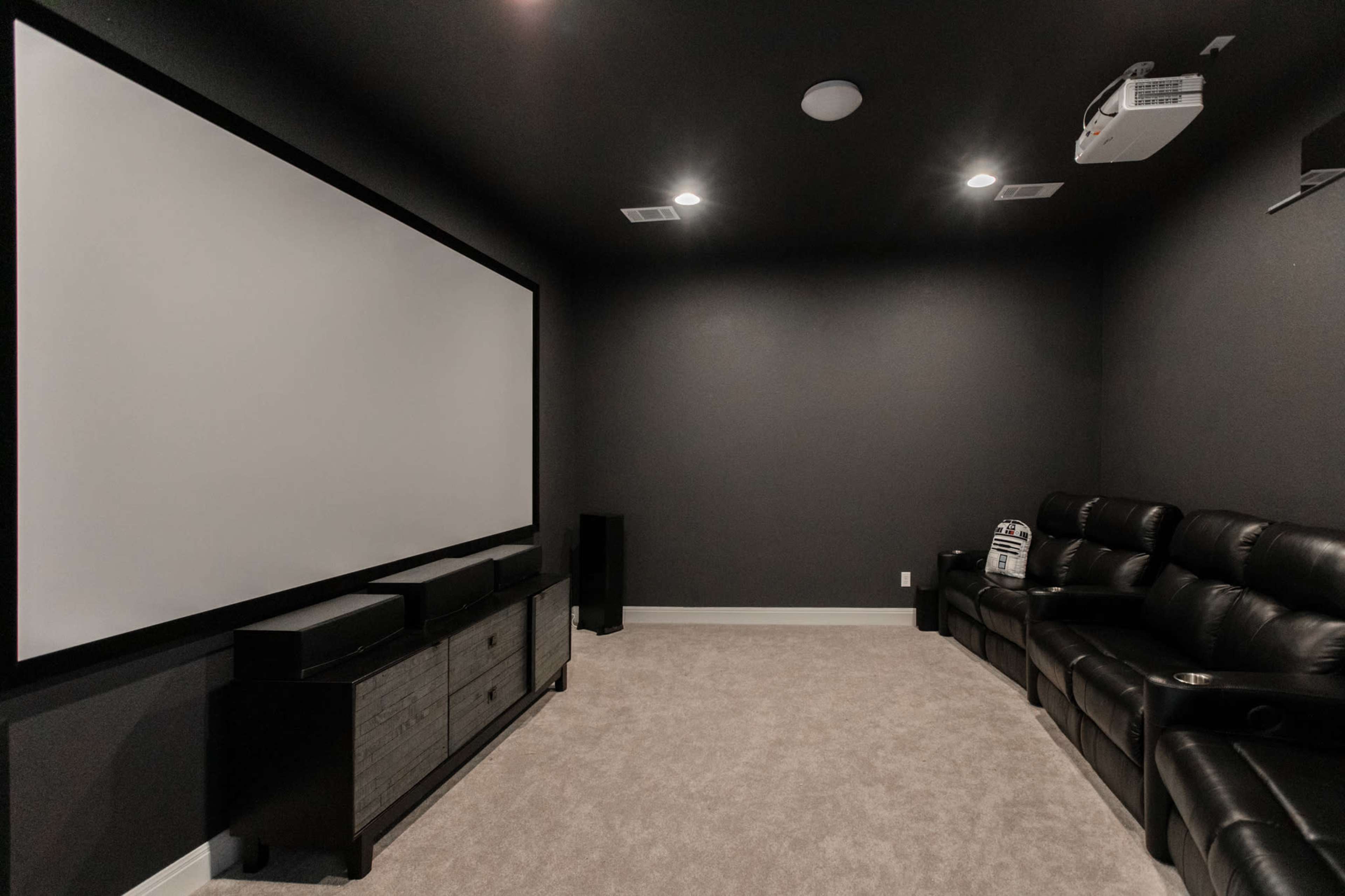 A dimly lit home theater features a large projection screen, a projector mounted on the ceiling, and a row of black leather reclining seats.