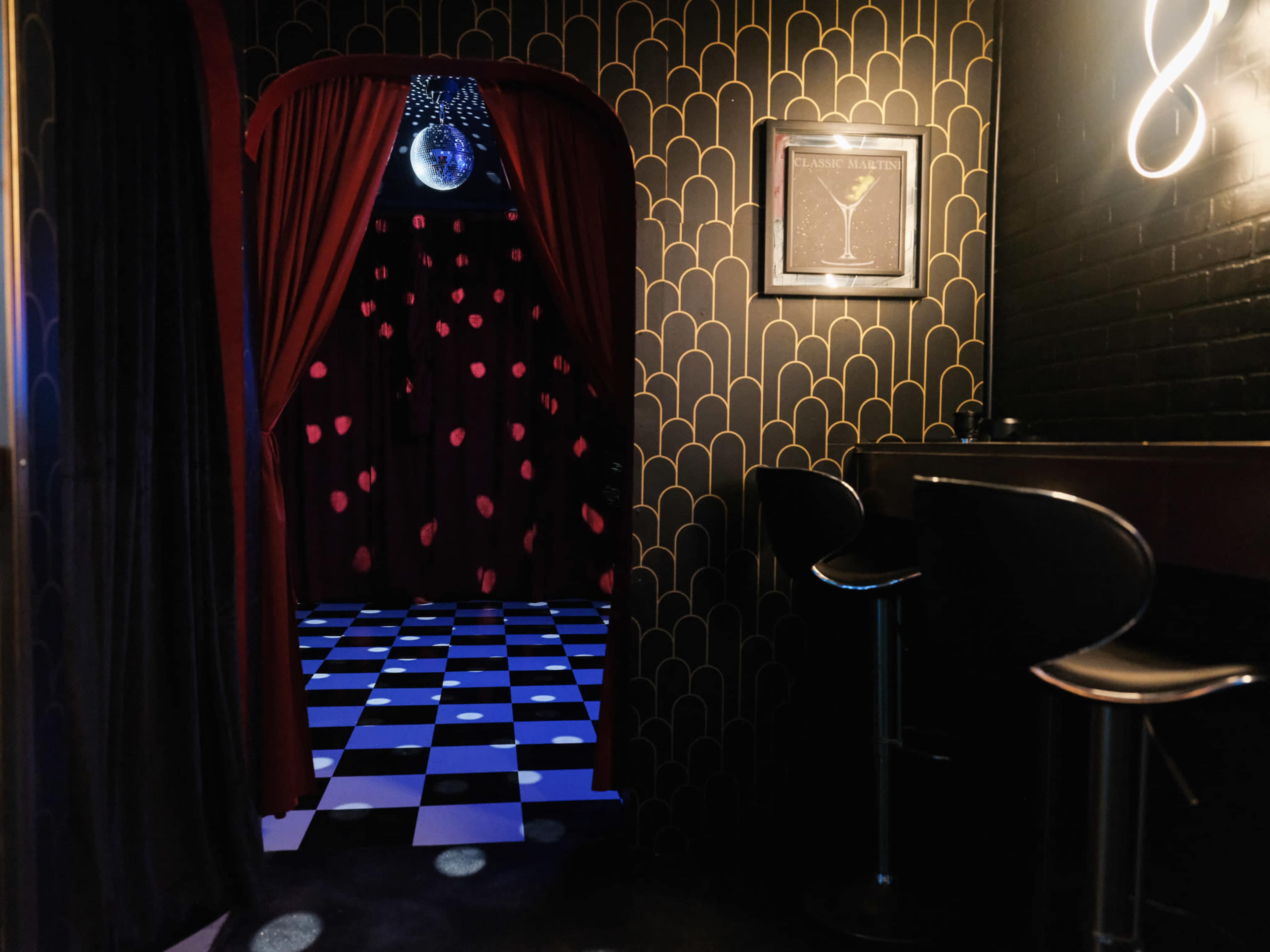 A dimly lit lounge features a black and white checkered floor and a doorway framed by red curtains, leading to a space adorned with heart-shaped decorations and a disco ball.