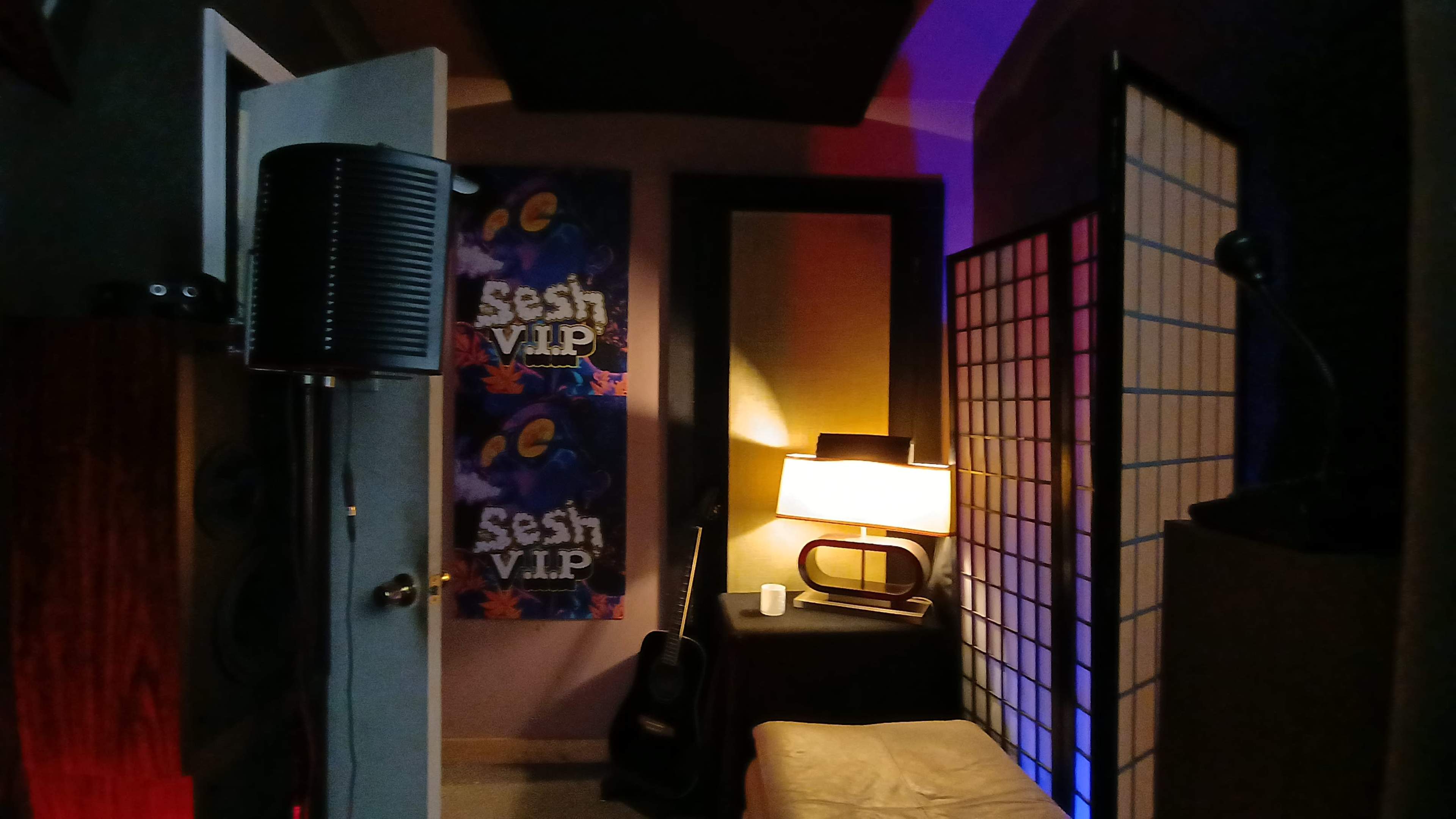 The image shows a small music studio with a microphone setup, a guitar leaning against a wall, a lamp, and decorative panels.
