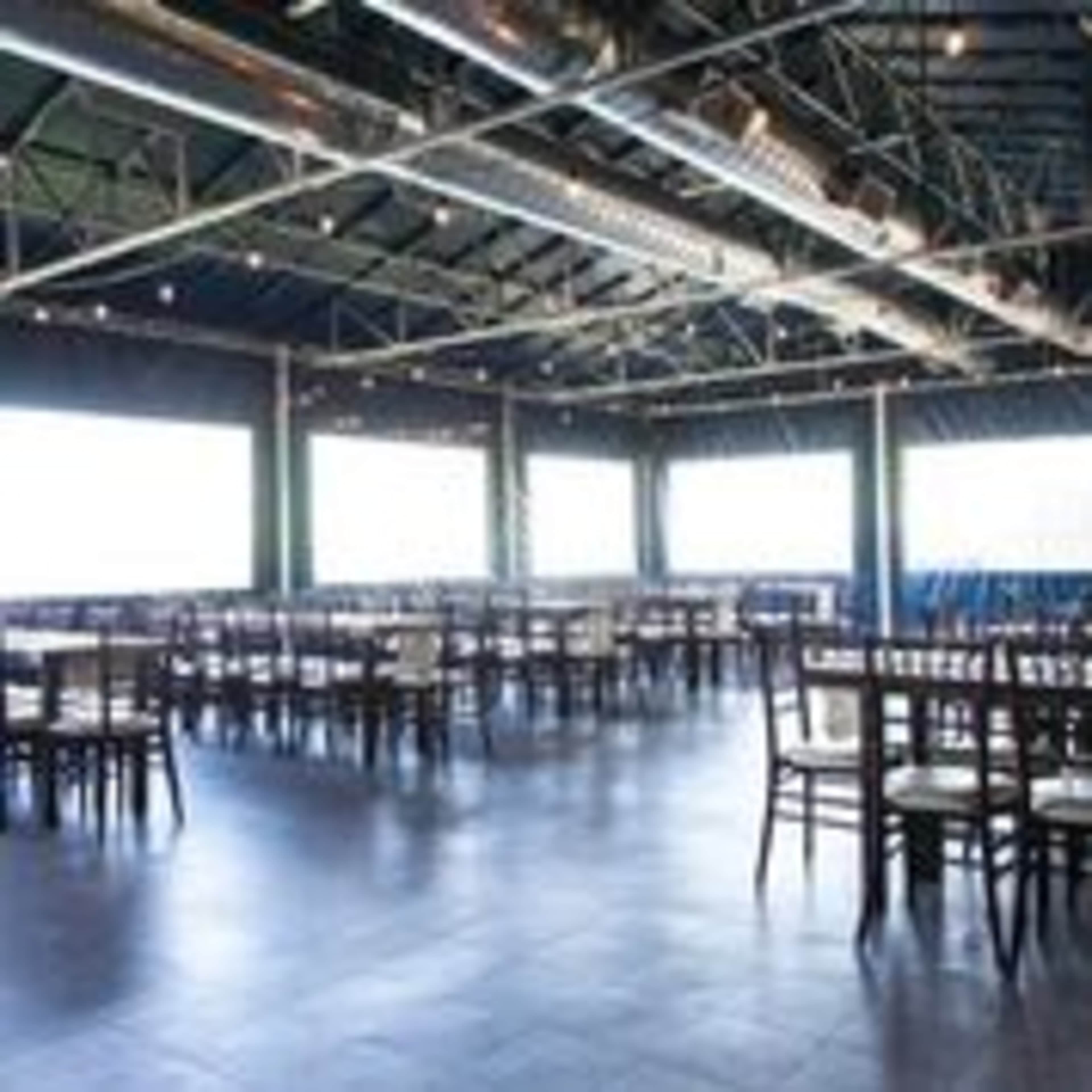 An empty restaurant interior with rows of wooden tables and chairs, large windows, and exposed metal beams.