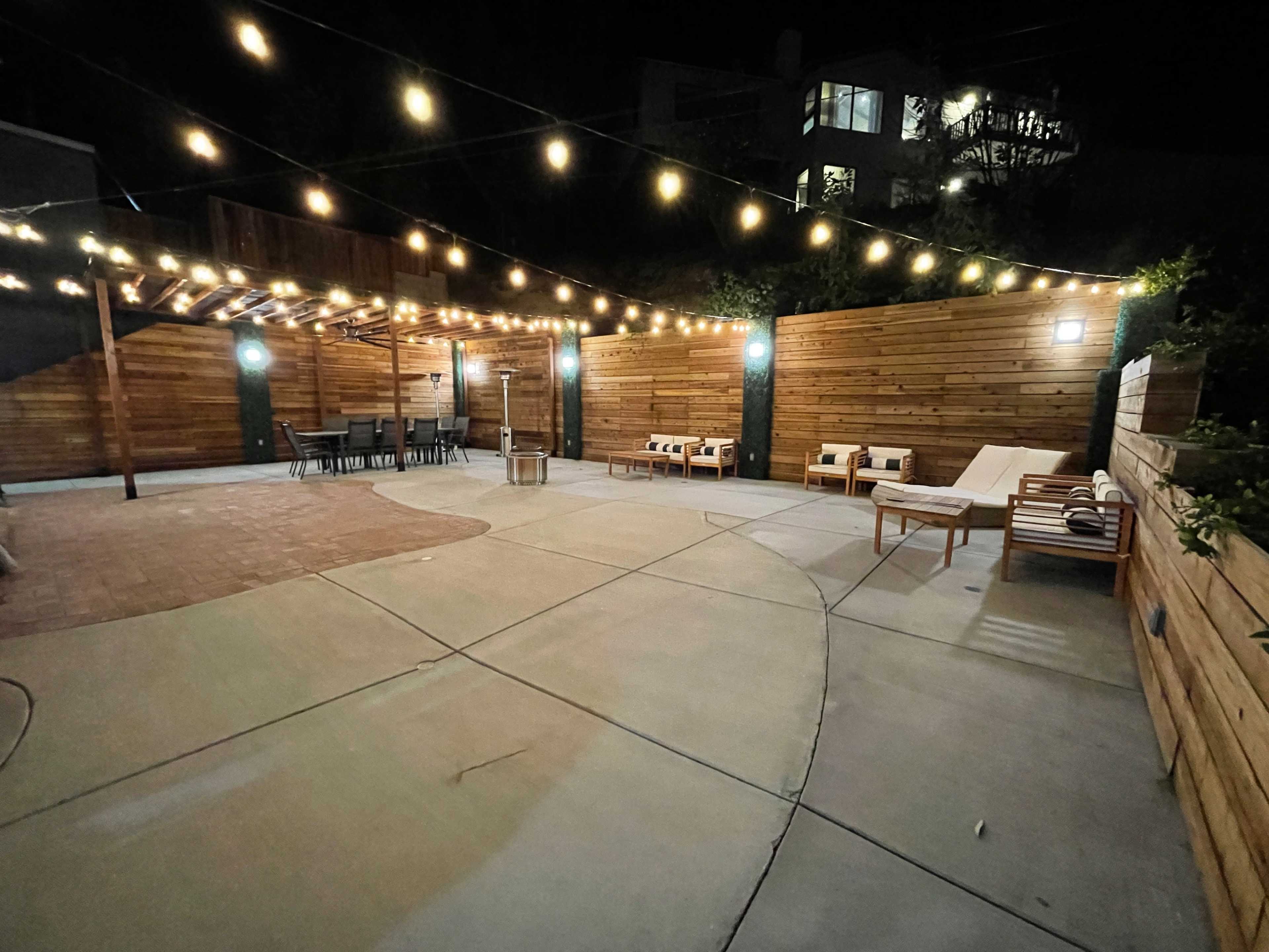 A patio area at night, illuminated by string lights, featuring wooden walls, seating, and a paved surface.