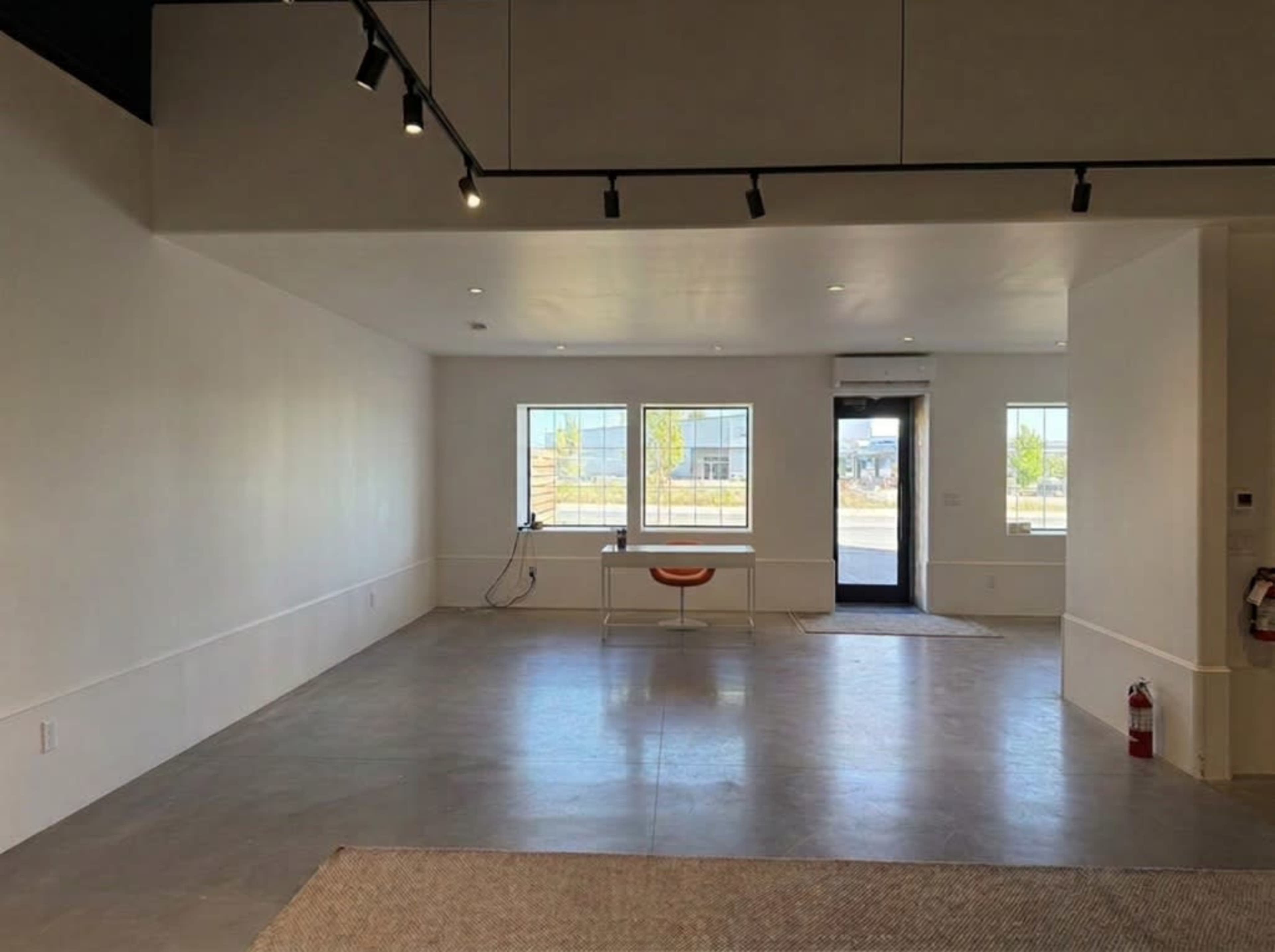 The image shows an empty interior space with gray concrete flooring, white walls, and large windows allowing natural light.