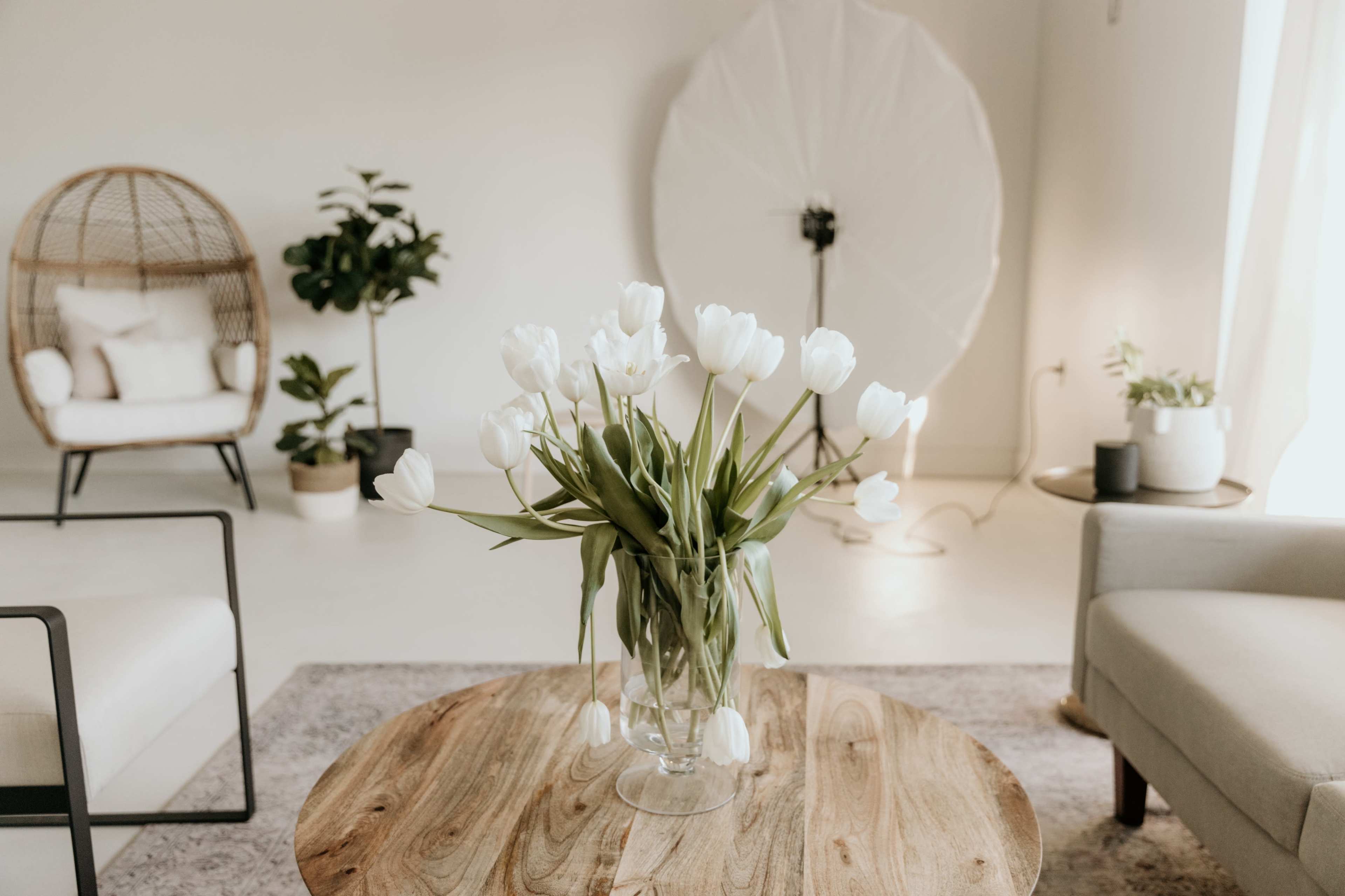 A vase of white tulips sits on a wooden table in a bright, uncluttered interior featuring a plant and a comfortable seating area.