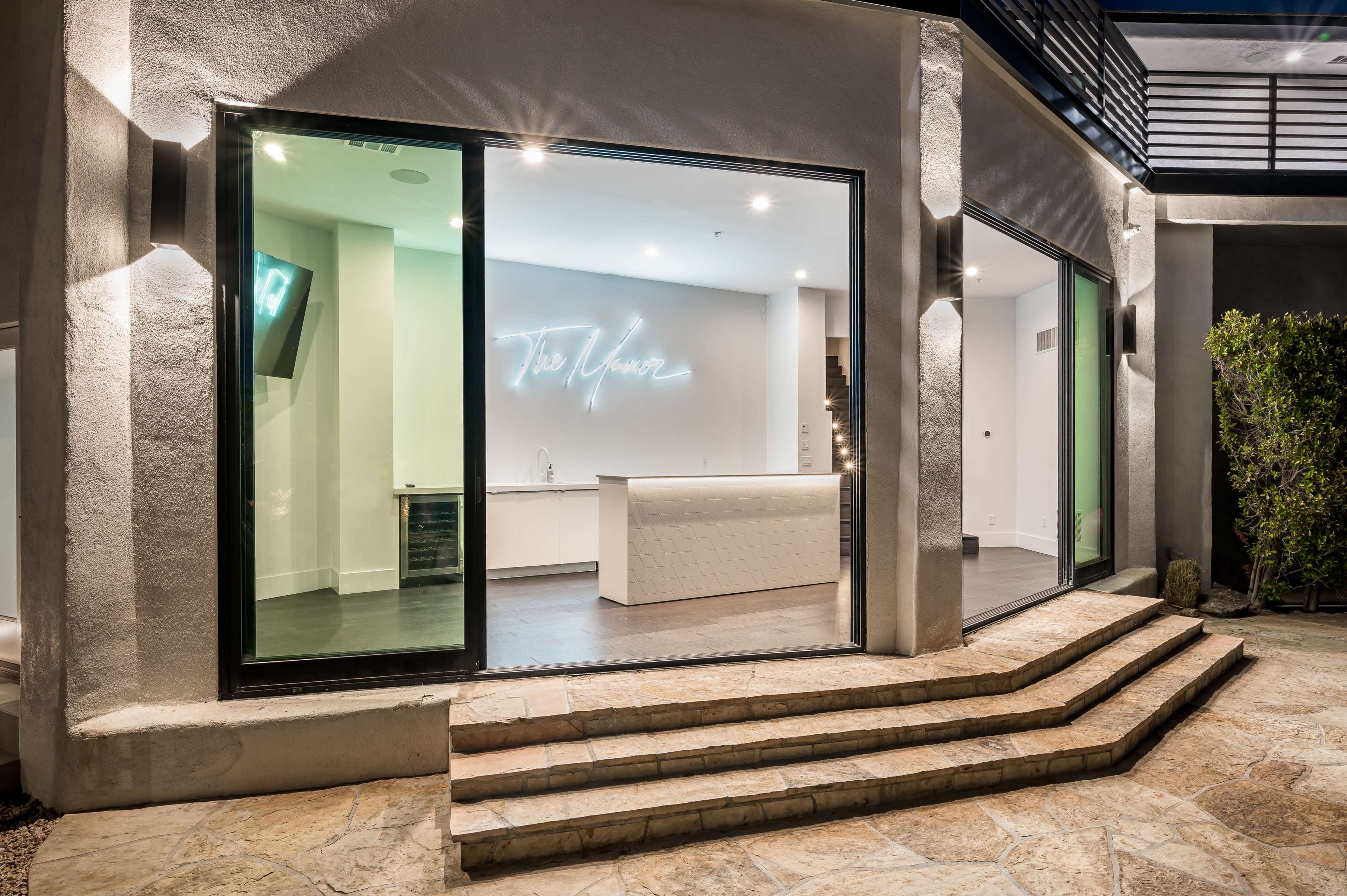A modern home exterior featuring large glass doors that open to a sleek white kitchen with a neon sign.