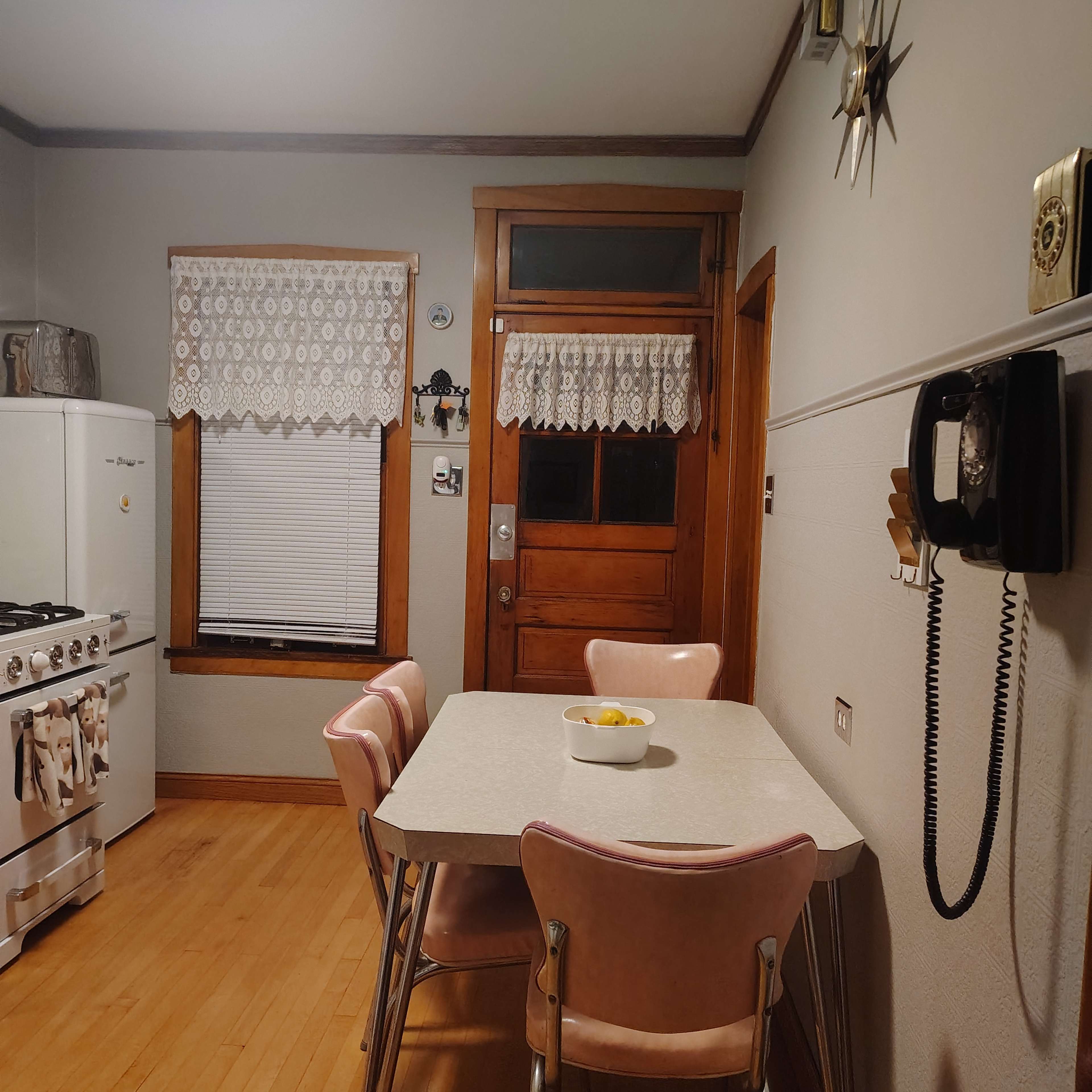 The image shows a simple kitchen with a table and pink chairs, a stove, and a door leading outside.