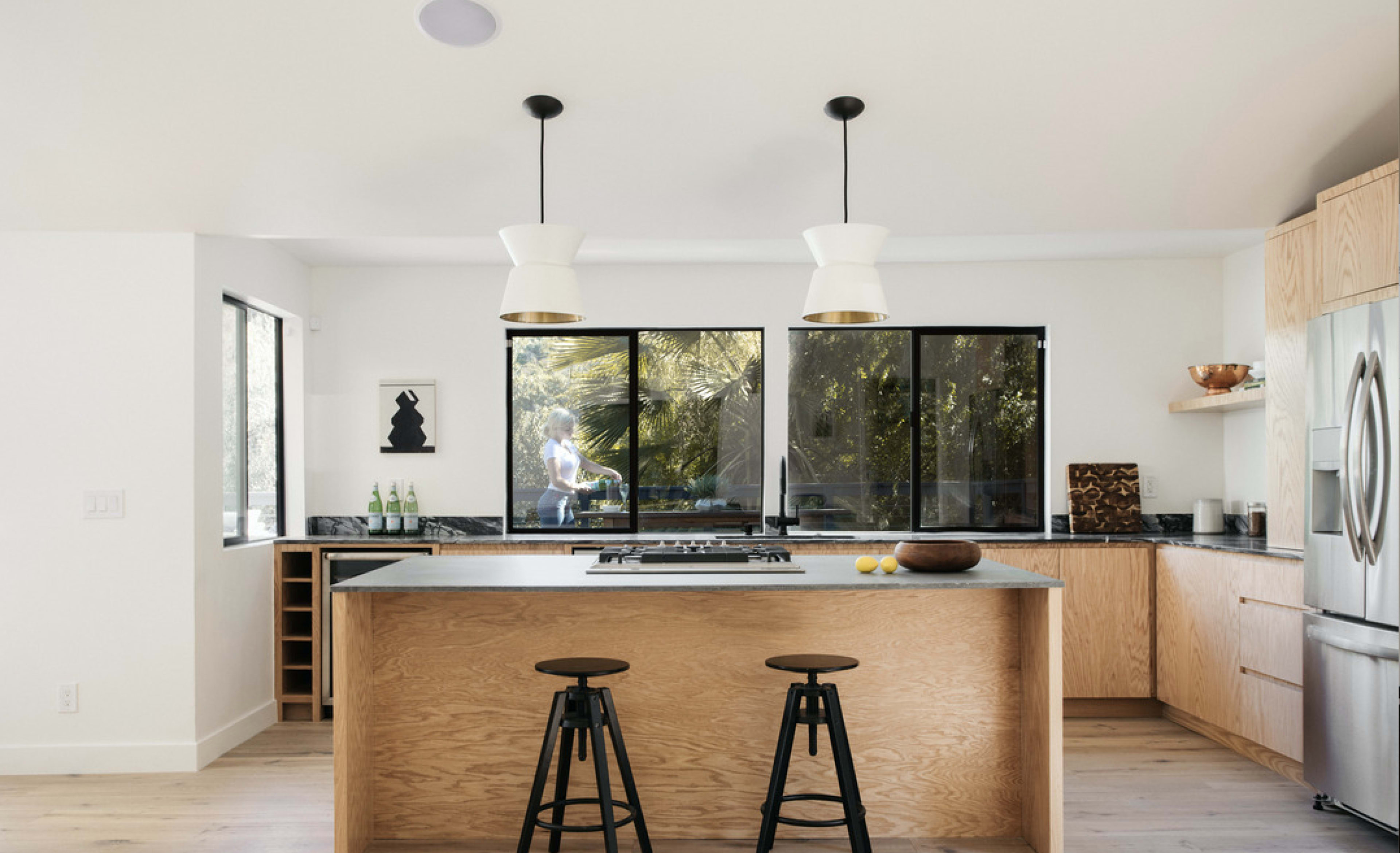 A modern kitchen features a central island with two bar stools, large windows showcasing greenery, and sleek cabinetry.