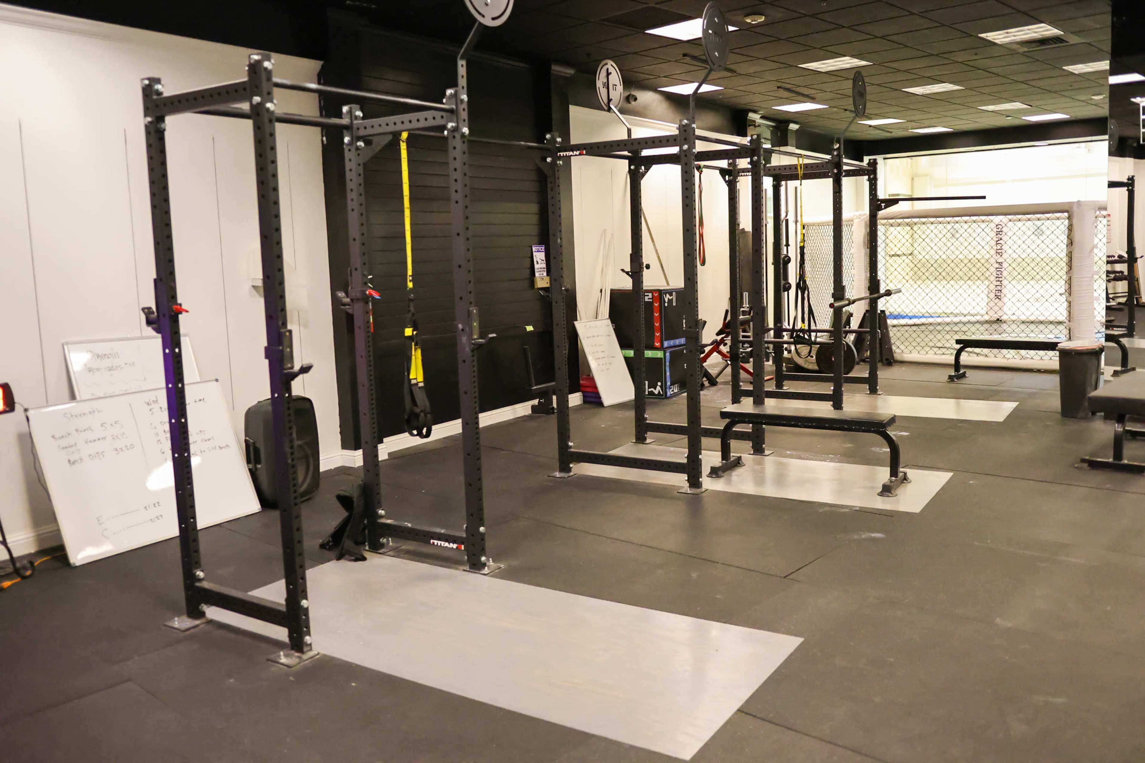The image shows a gym interior with multiple squat racks and exercise equipment on a rubber flooring surface.