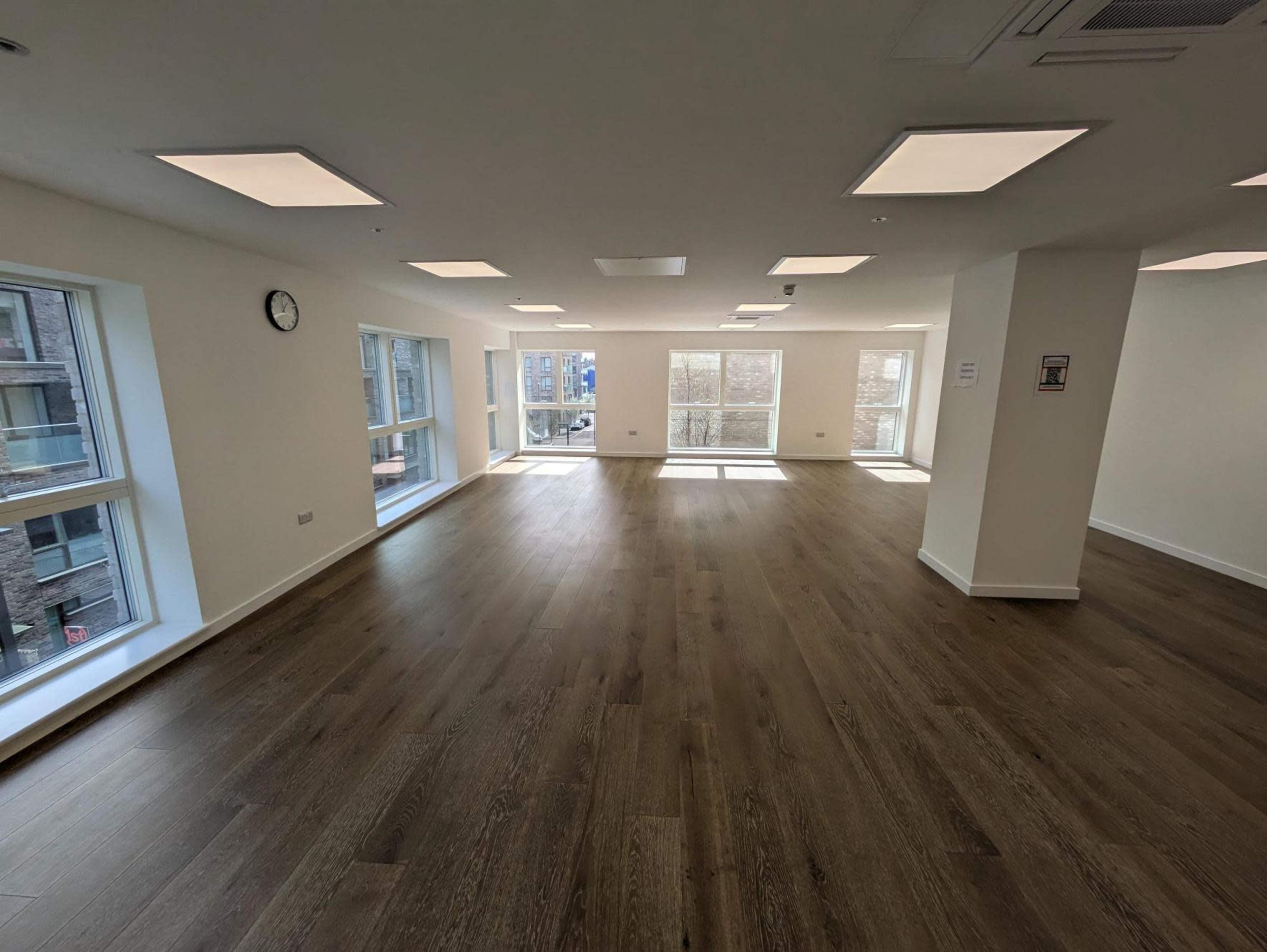 The image shows a spacious, well-lit empty office with large windows and wooden flooring.