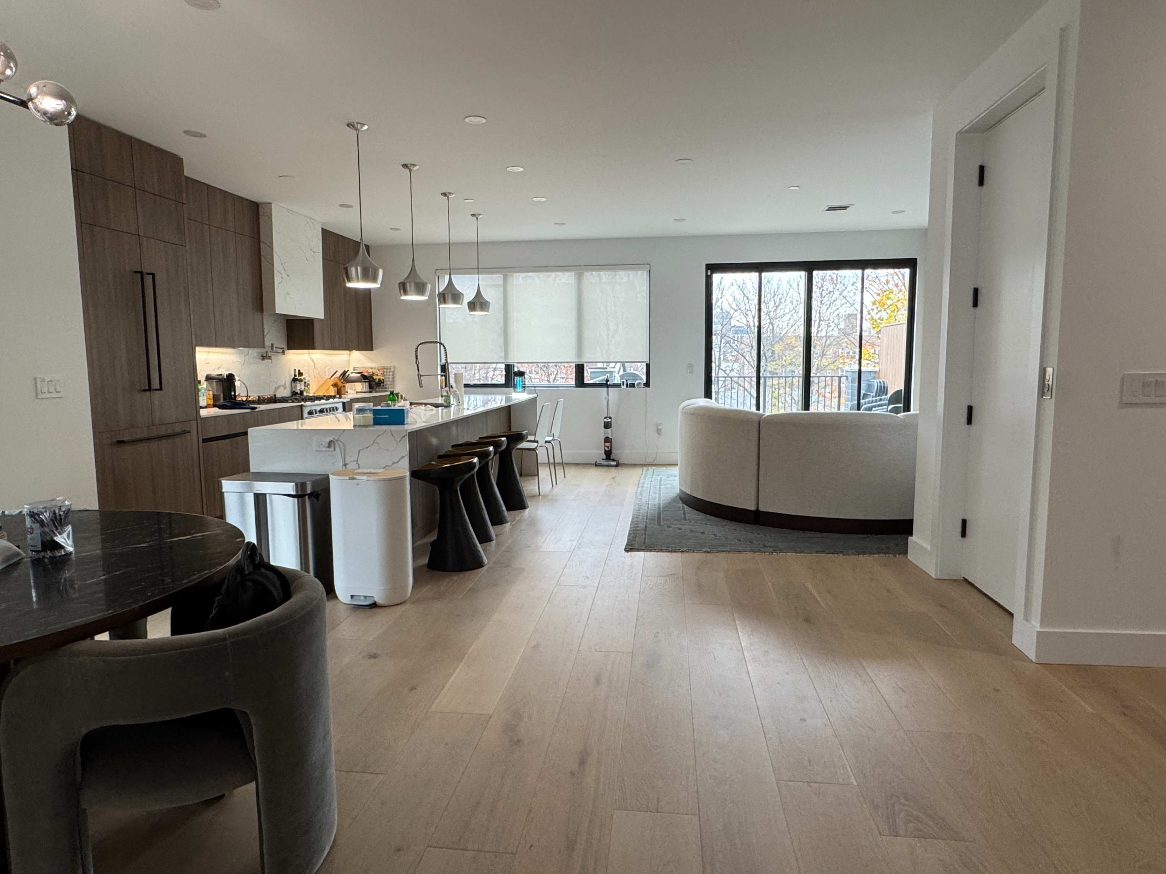 The image shows a modern kitchen and living area with an open layout, featuring a kitchen island, dining table, and seating area with large windows.