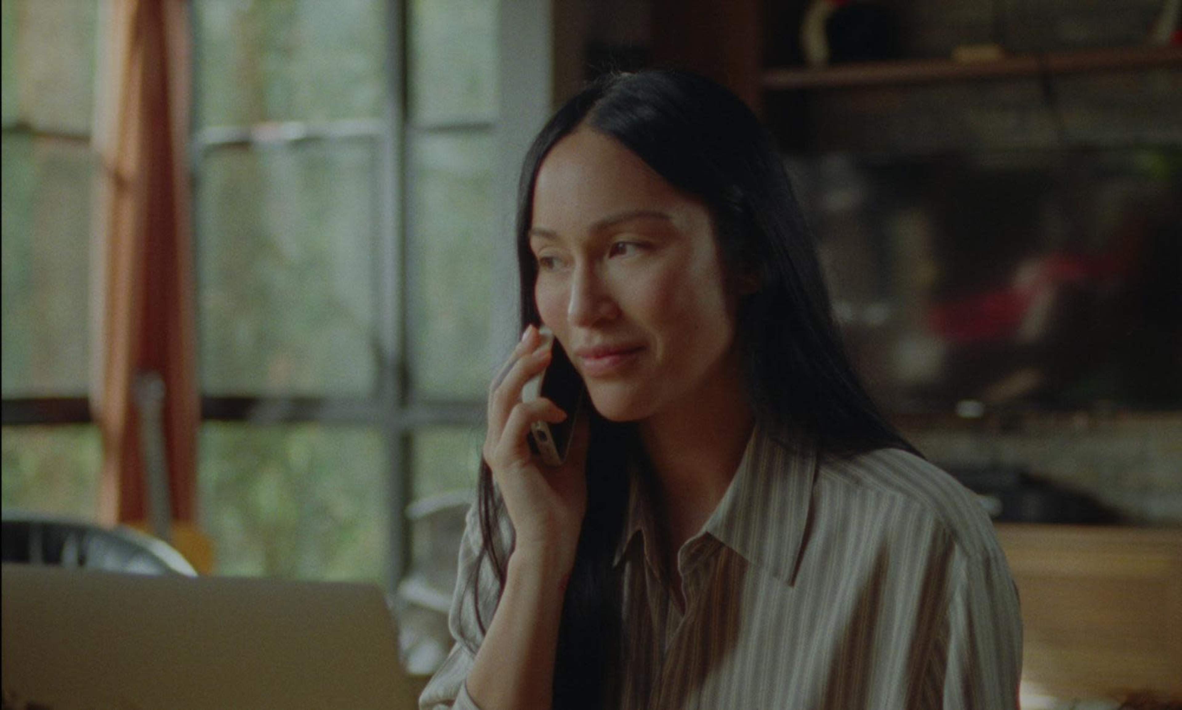 A woman with long dark hair is sitting at a desk, smiling while talking on a phone, with a laptop open in front of her and a window in the background.