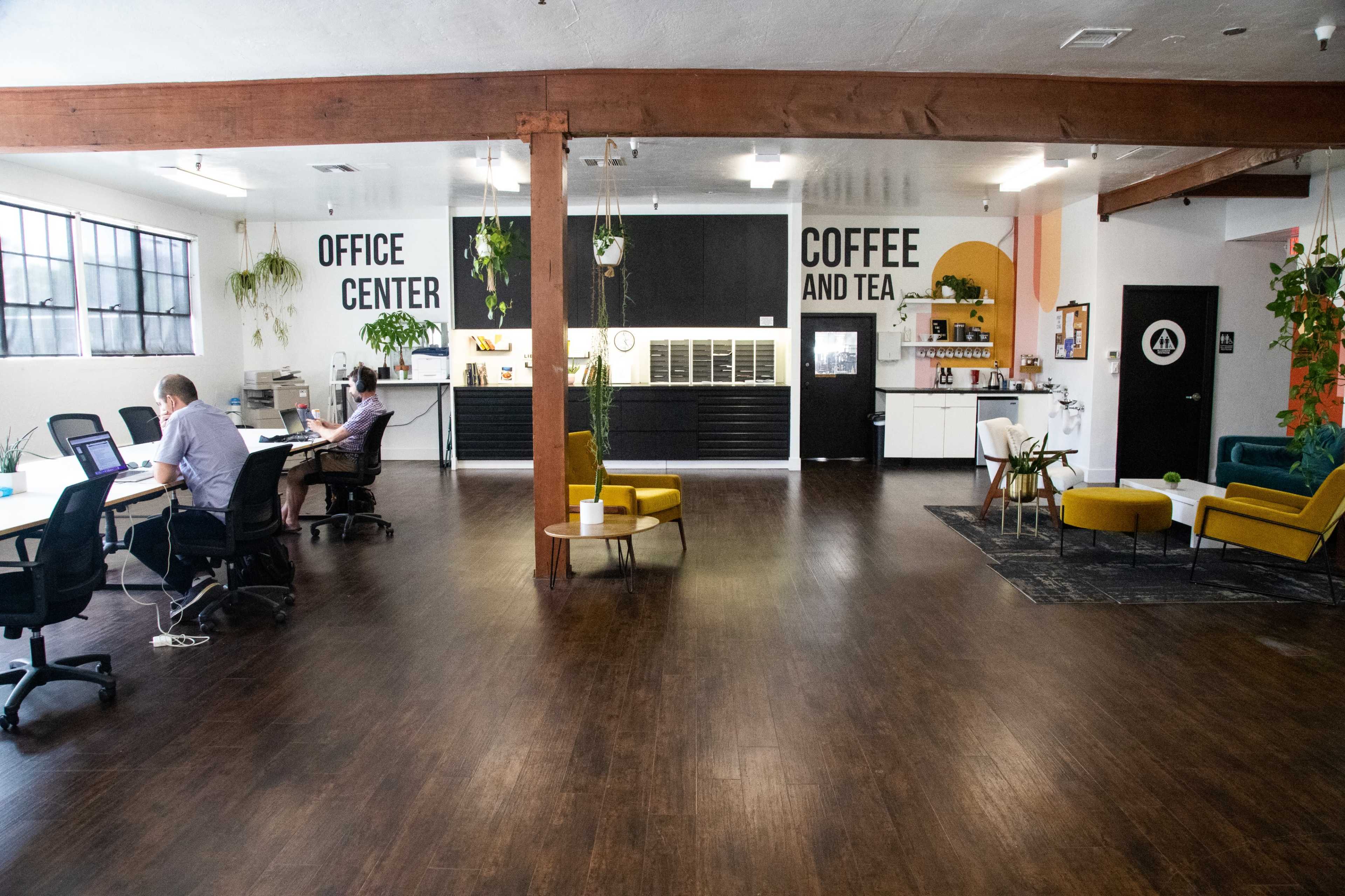 A modern office space featuring workstations, a coffee area, and ample greenery, with a prominent sign that reads "OFFICE CENTER" and "COFFEE AND TEA."