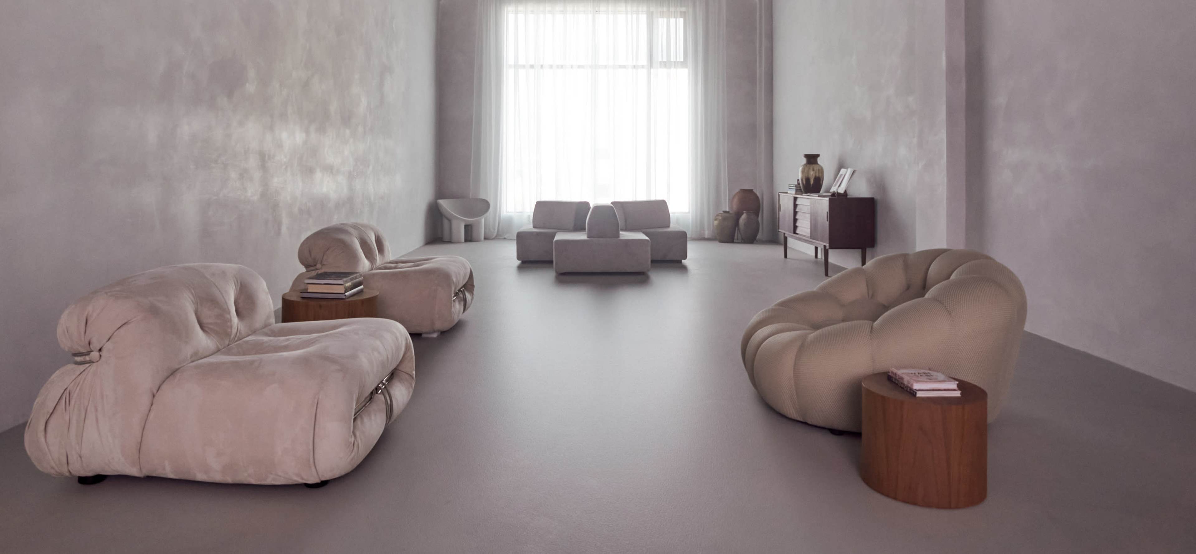 The image shows a minimalist interior featuring several contemporary sofas and a low coffee table, with large windows providing natural light.