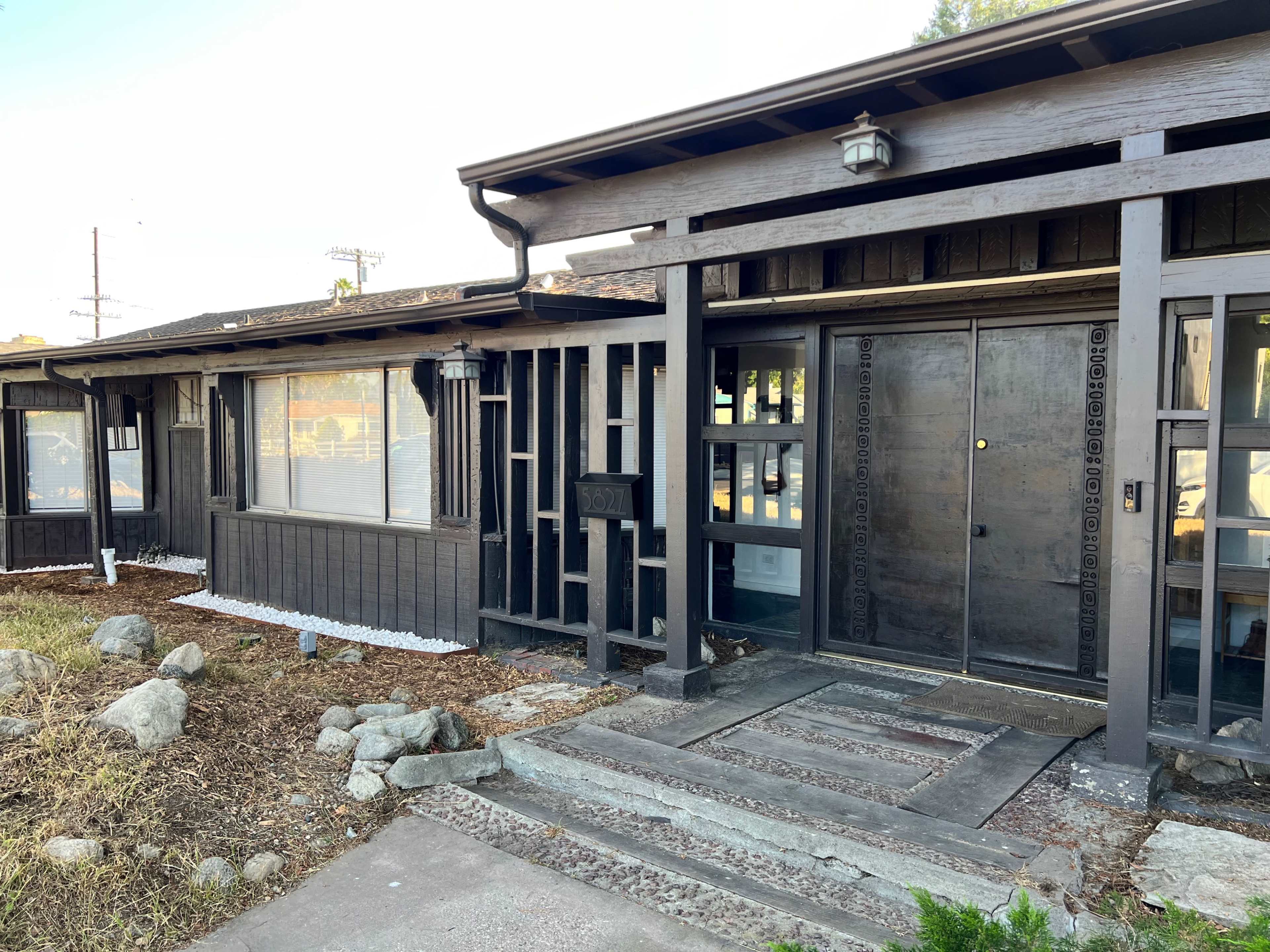 A single-story house with a dark wooden facade and a pathway leading to a large front door, surrounded by a rocky garden.