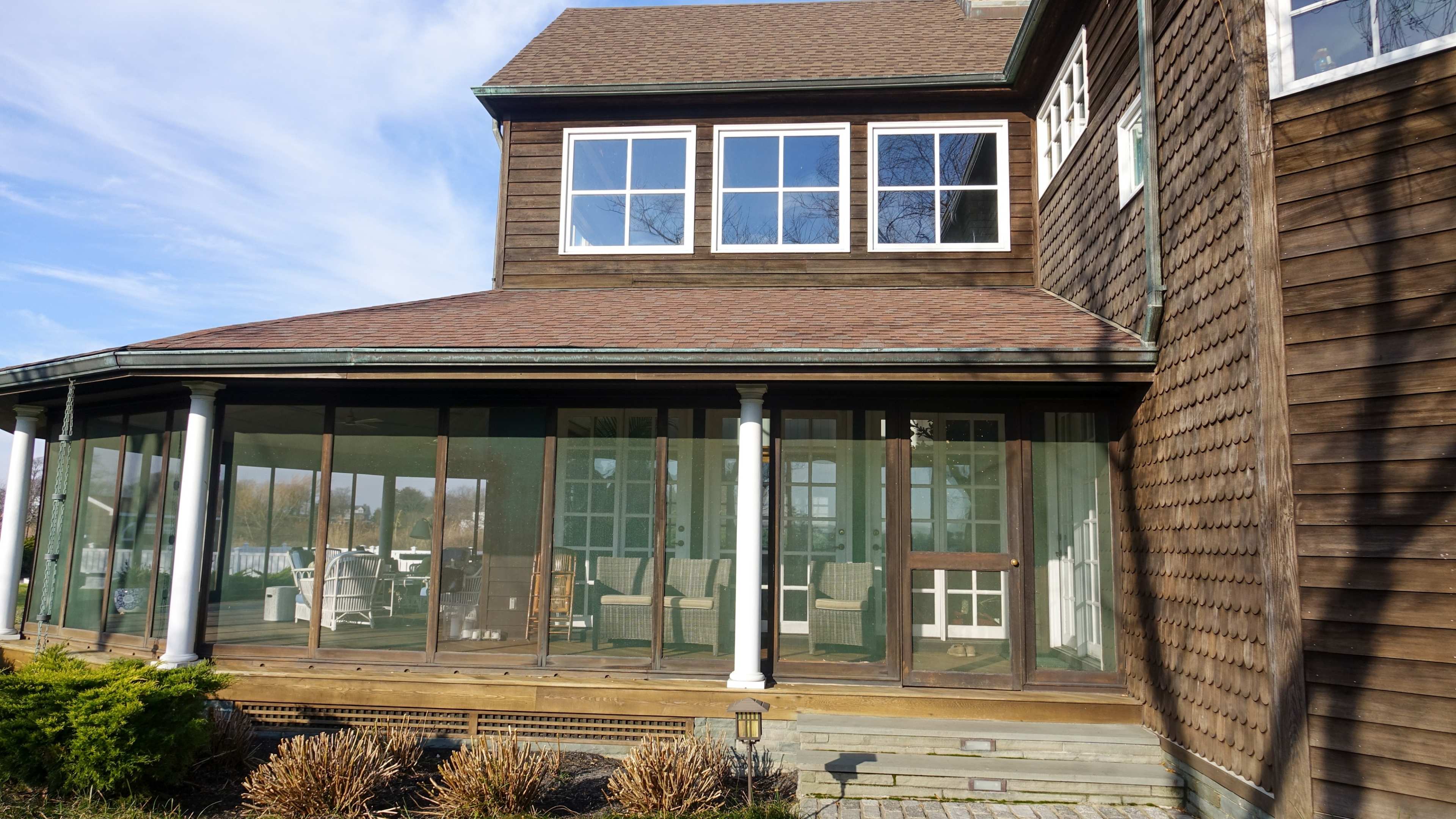 A wooden house with a screened-in porch featuring large windows and a view of surrounding greenery.