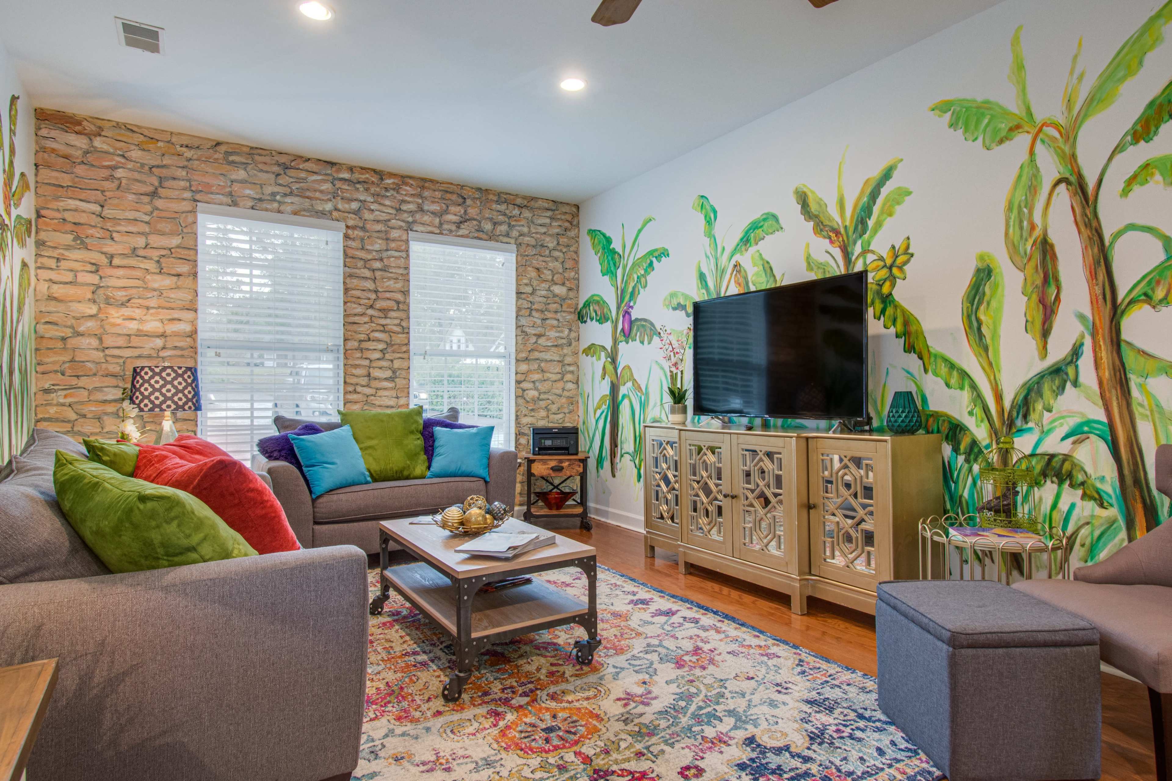 A living room features a stone accent wall, colorful sofa cushions, a wooden coffee table, and a television against a backdrop of tropical wall murals.